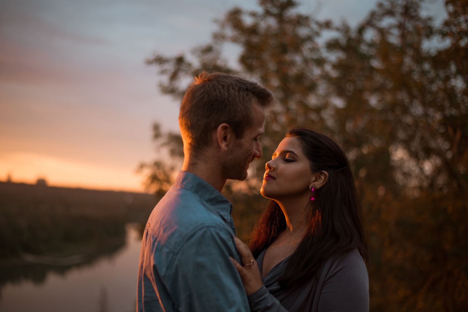 A romantic couple embraces during a warm sunset by a tranquil river, bathed in golden light.