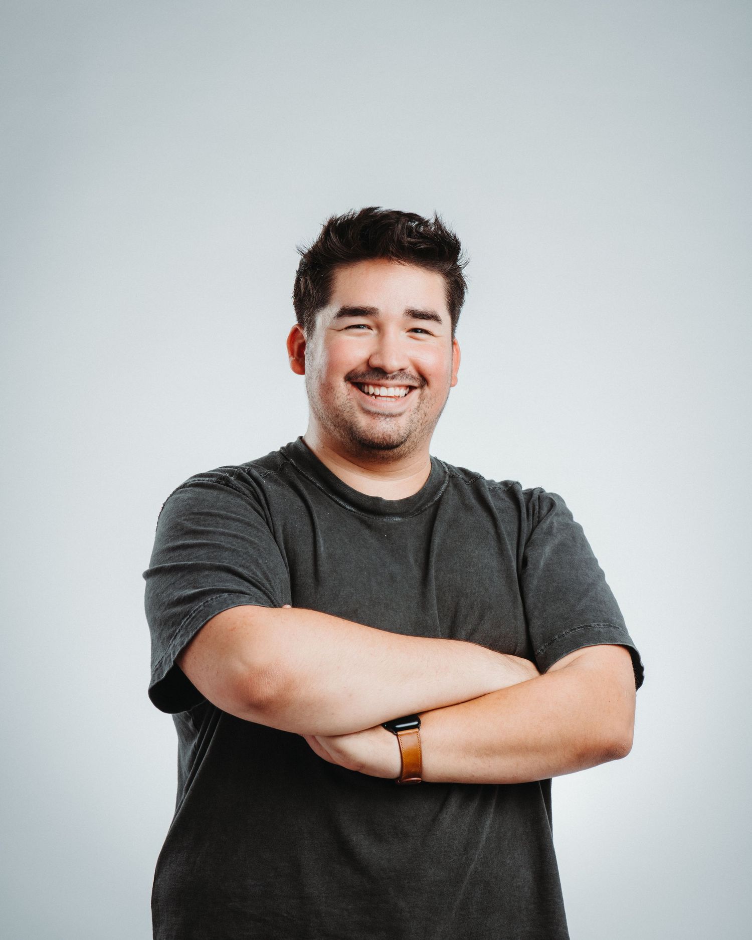 Person wearing dark grey shirt stands with crossed arms against white background while smiling warmly.