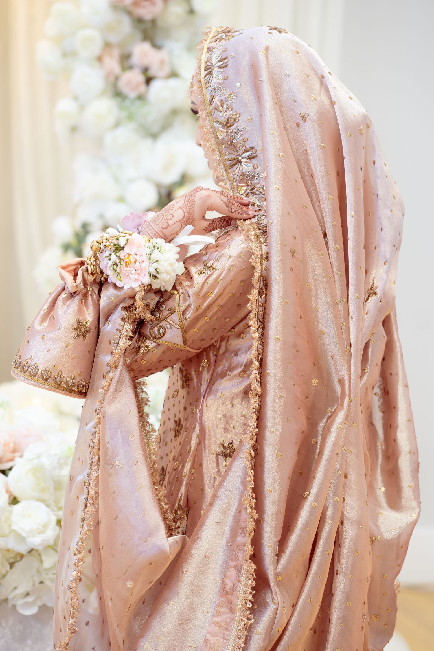 A bride in an elegant rose gold embroidered sari and veil holds a delicate floral bouquet at a wedding ceremony.