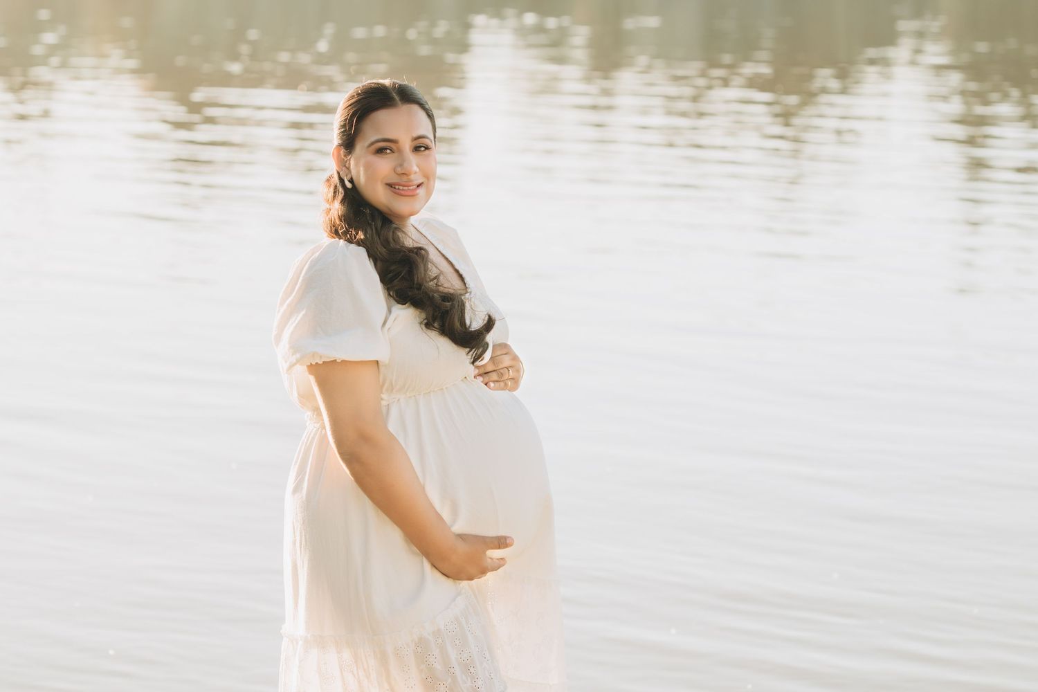Smiling pregnant woman in white dress cradling baby bump near serene water in soft natural light.