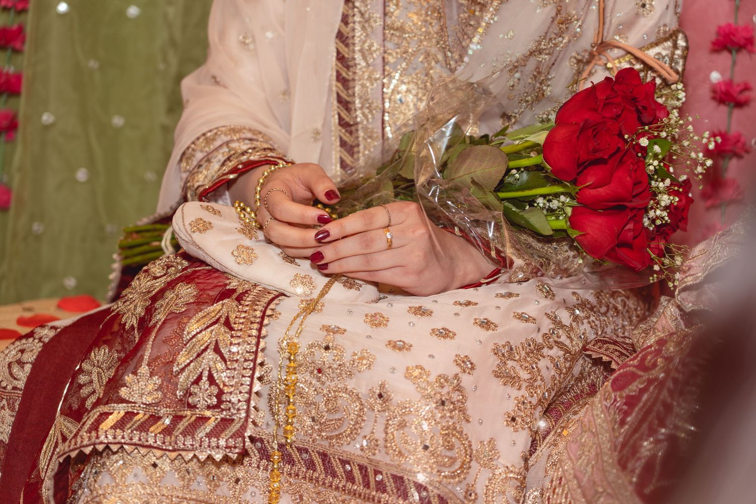 Bride in embroidered ivory and gold lehenga holding red roses bouquet with mehndi hands and gold bangles.