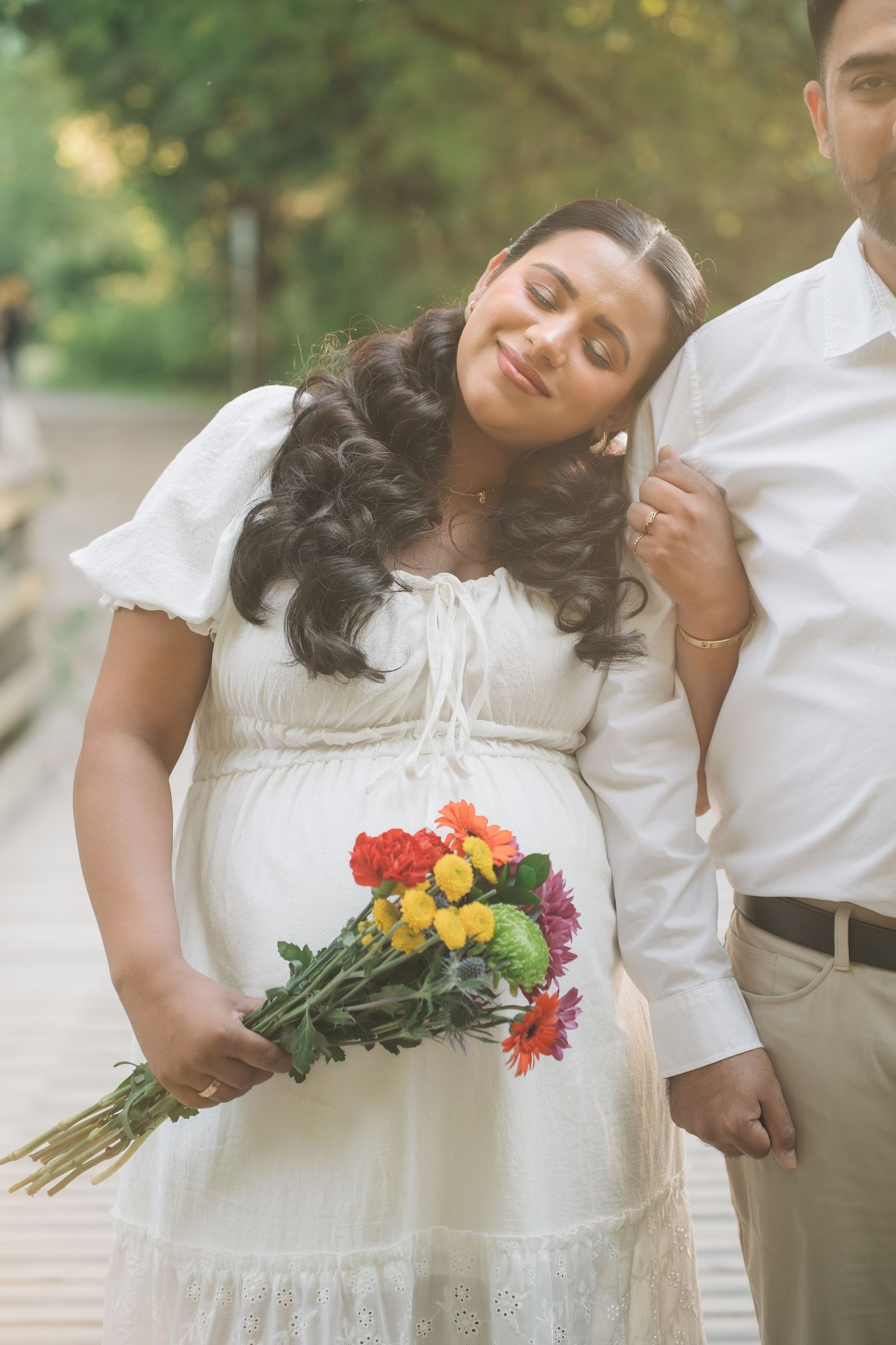 Pregnant woman in white dress holds colorful flowers, leaning on partner outdoors in a maternity photo shoot.