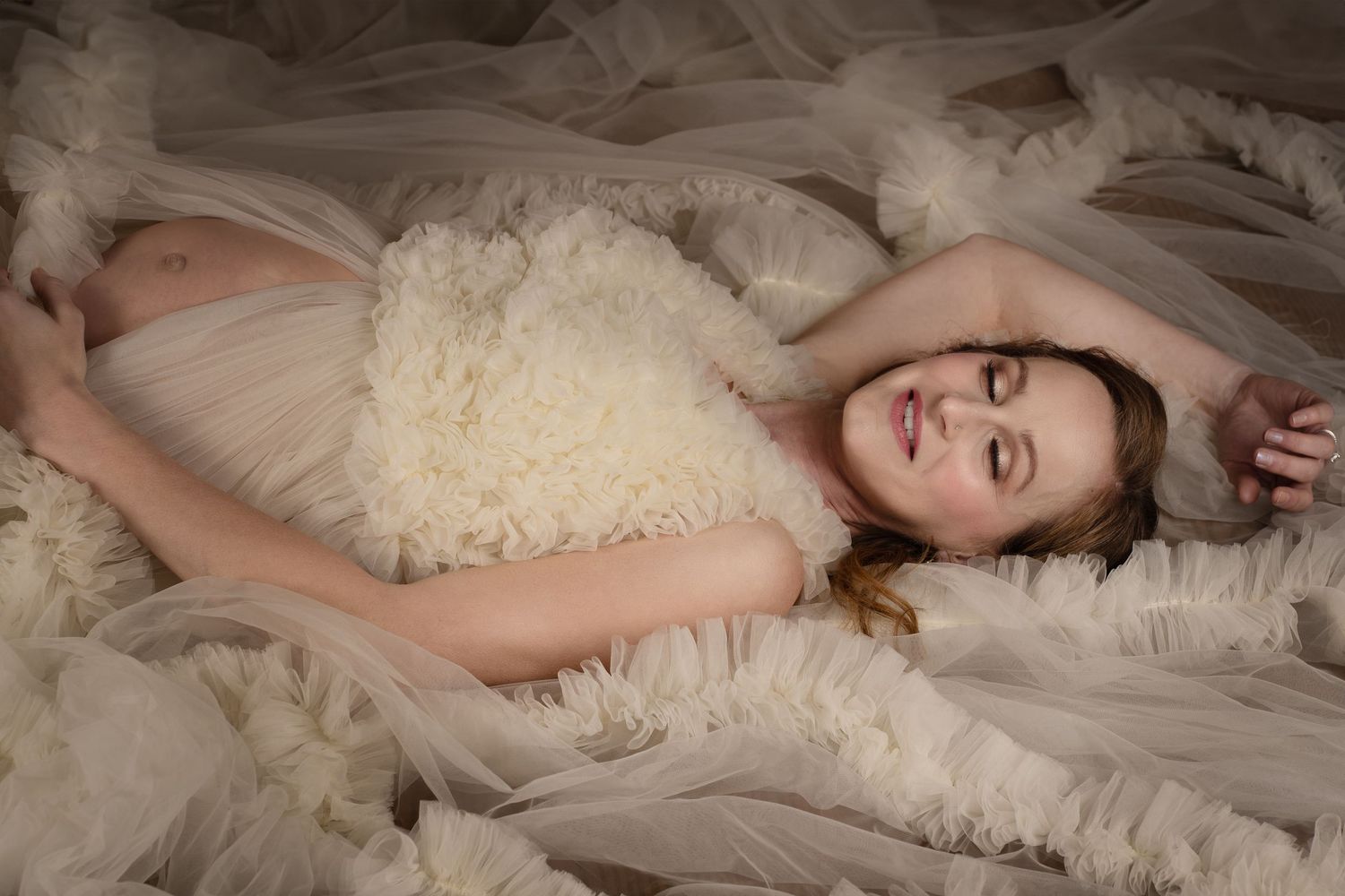 Pregnant woman in white ruffled dress lying gracefully on flowing tulle fabric in a dreamy maternity portrait.