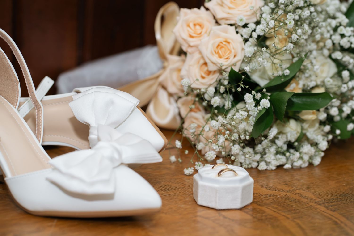 White bridal heels with bow detail beside a ring box and a bouquet of peach roses and baby's breath on wood.