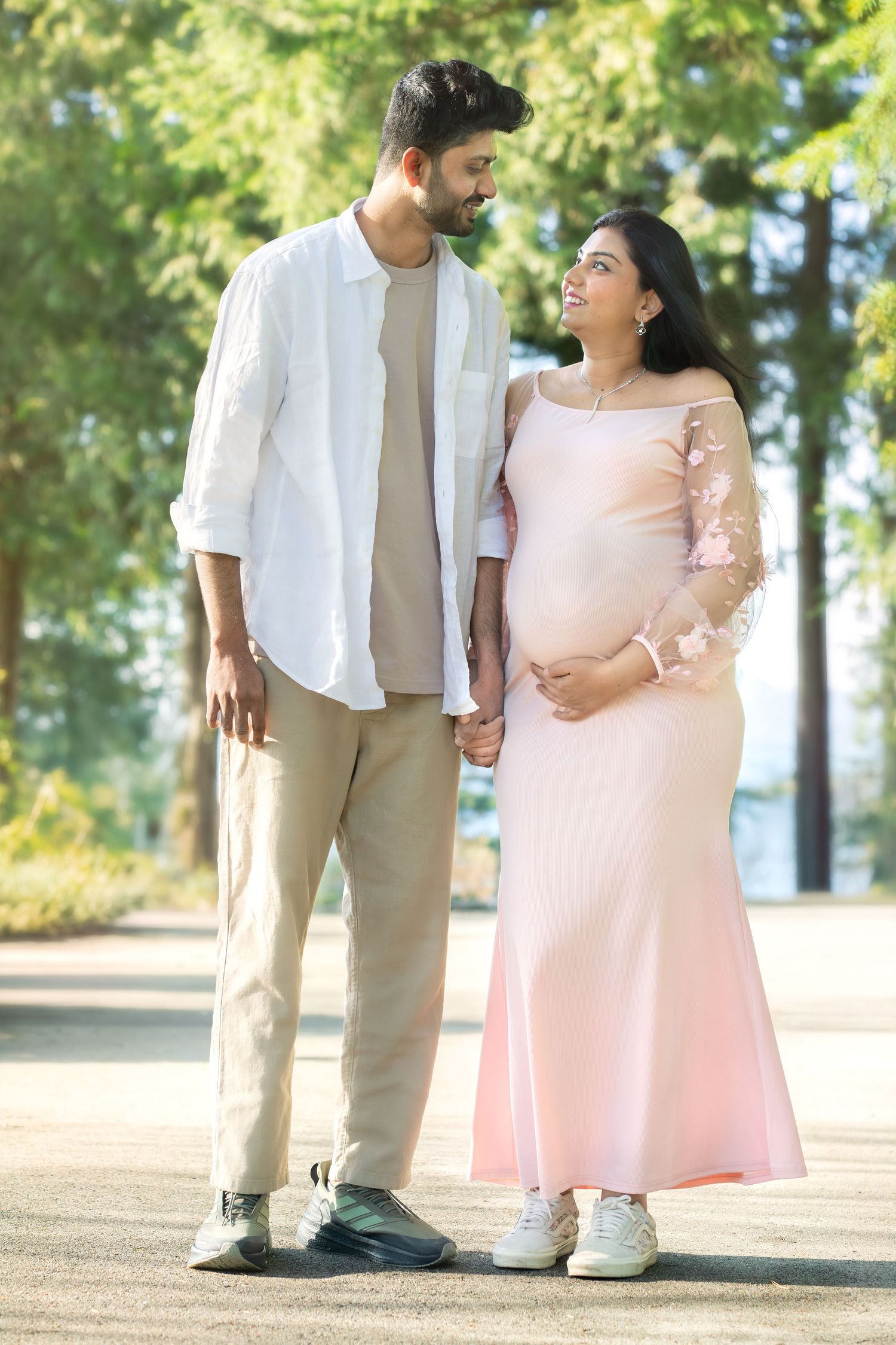 A pregnant Indian couple holds hands and smiles at each other outdoors, she wears a pink gown, he wears white shirt.