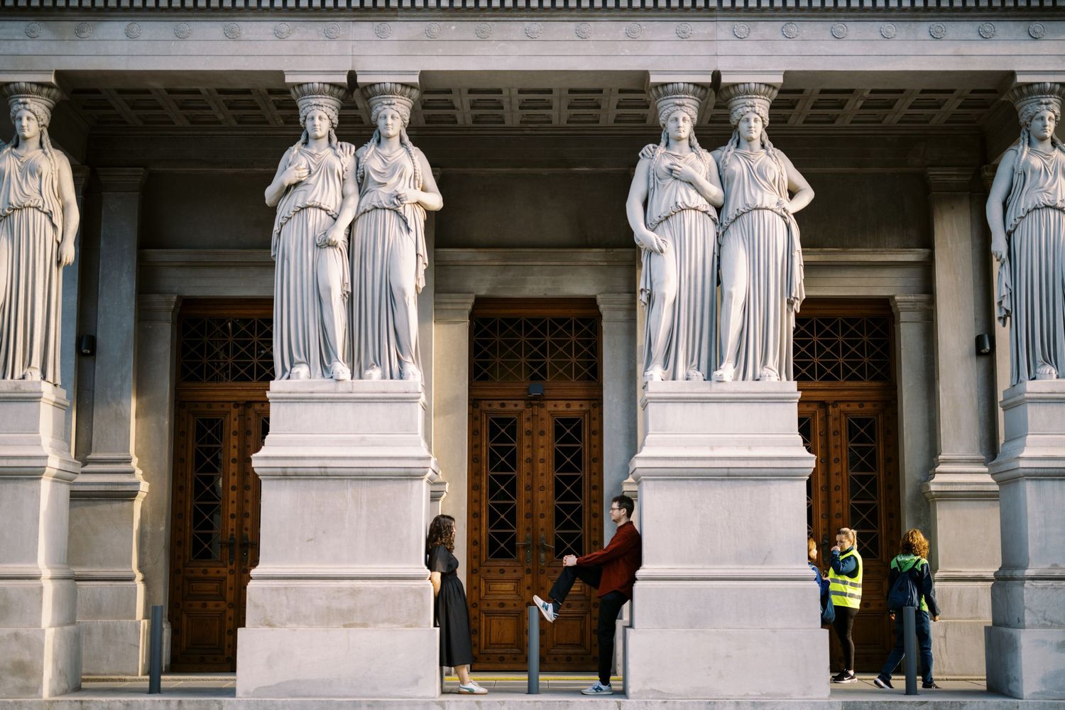 Classical stone statues flank ornate wooden doors on a grand building entrance with carved architectural details.