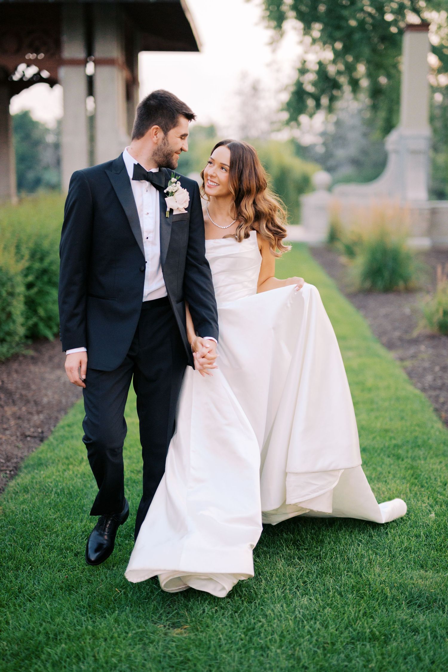 A couple in wedding attire walks together on a lush green lawn during an outdoor wedding photoshoot.