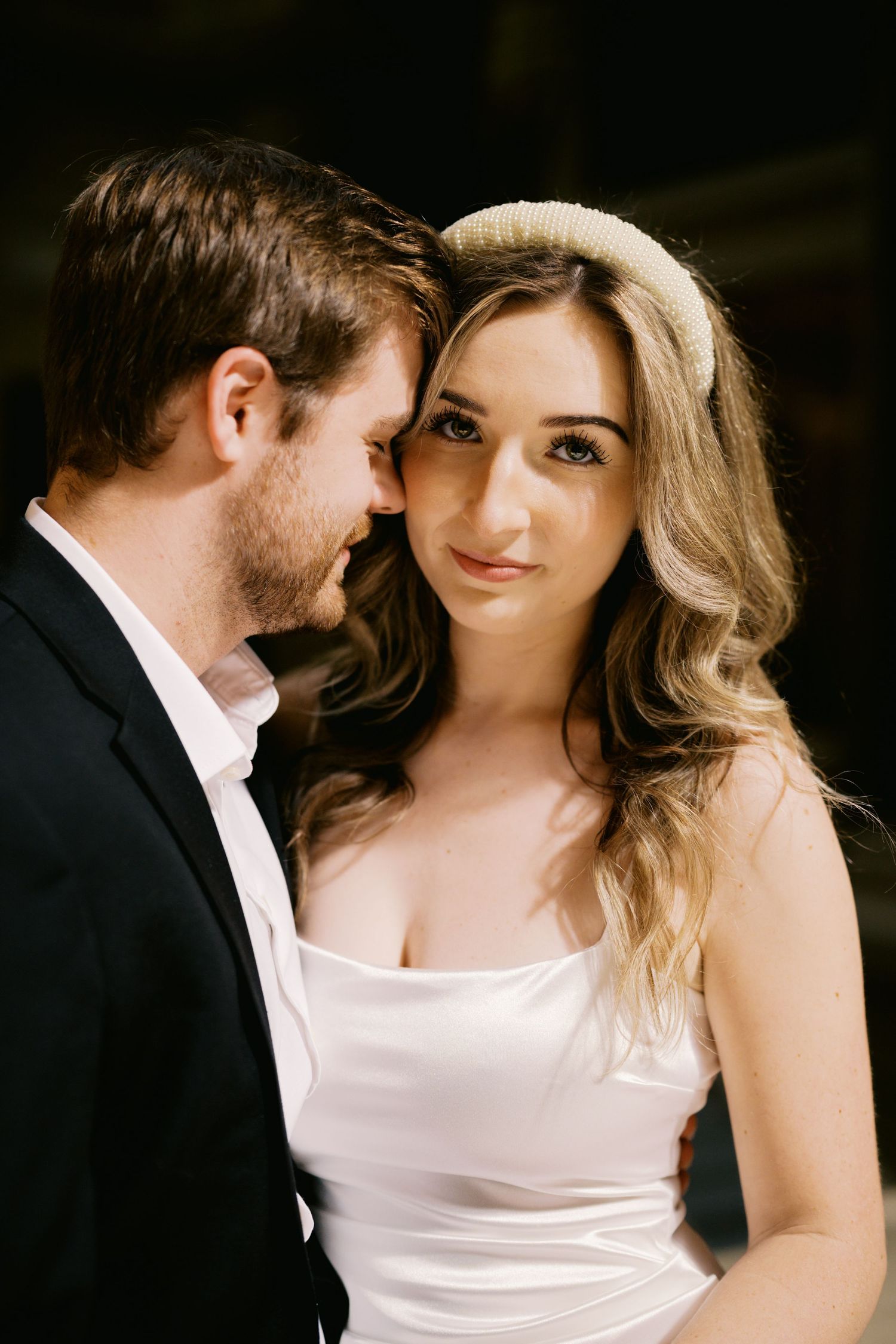 Romantic close-up portrait of a couple in formal attire with the woman wearing a white gown and headband.