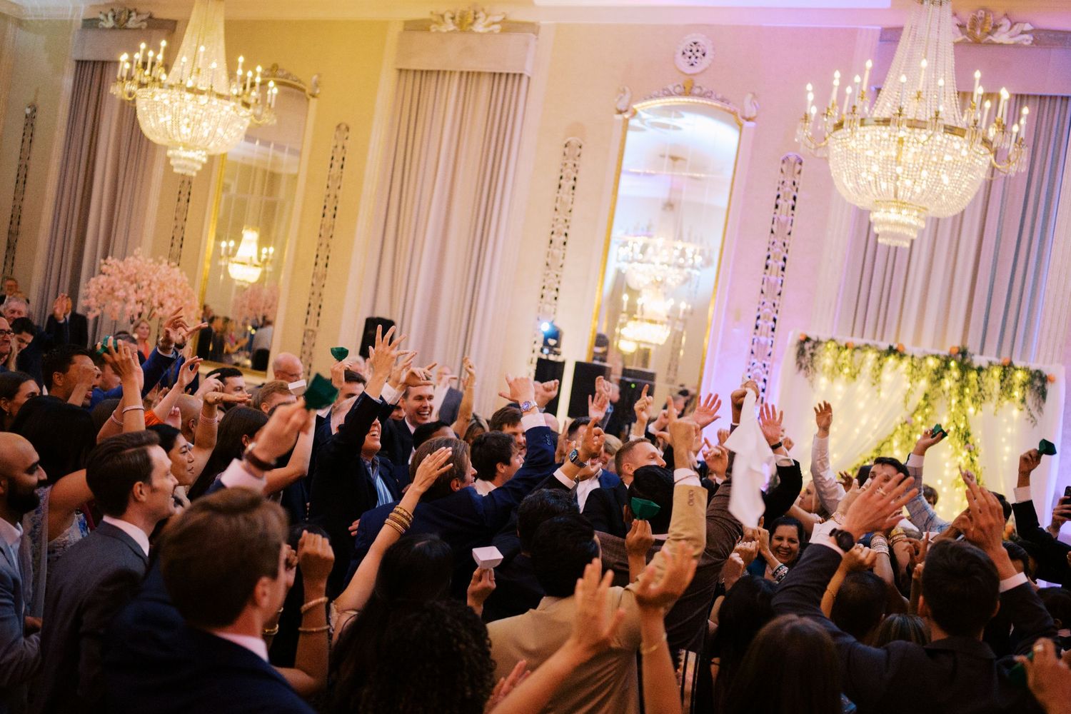 Wedding guests raise their hands in celebration at an elegant ballroom reception with crystal chandeliers and pink lighting.
