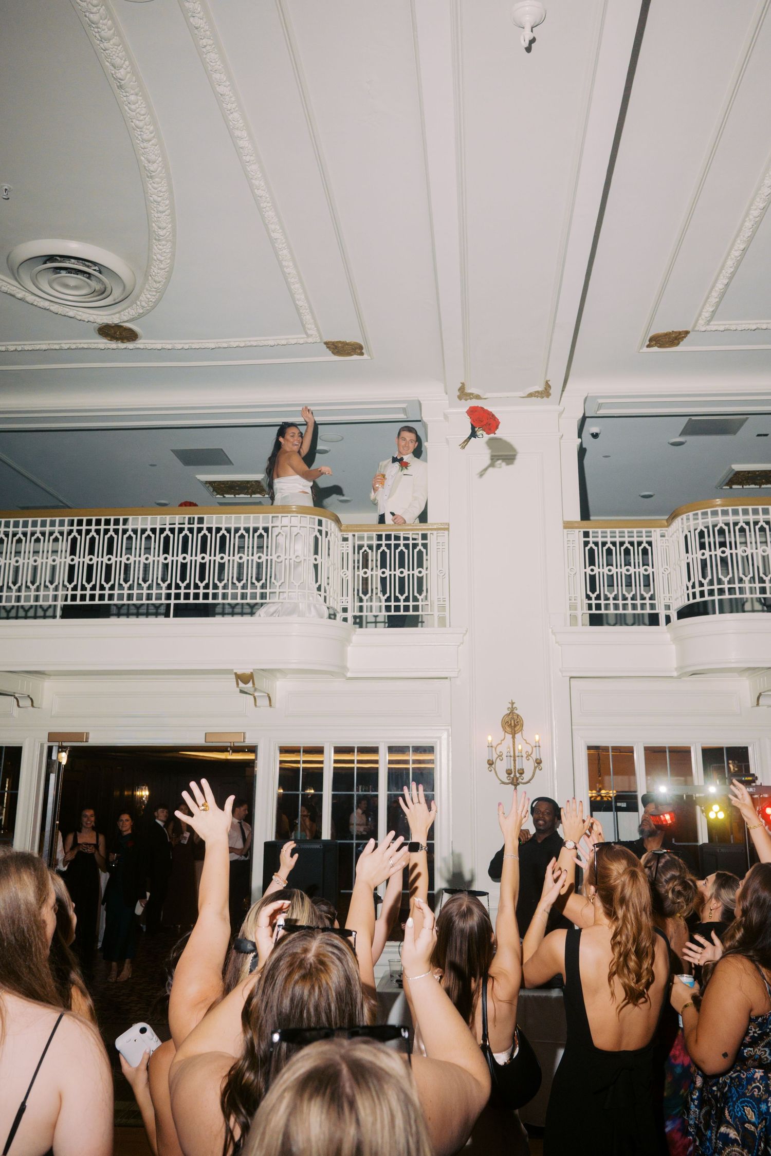 A crowd of wedding guests dancing with raised hands at a reception under an ornate white ceiling with chandeliers.