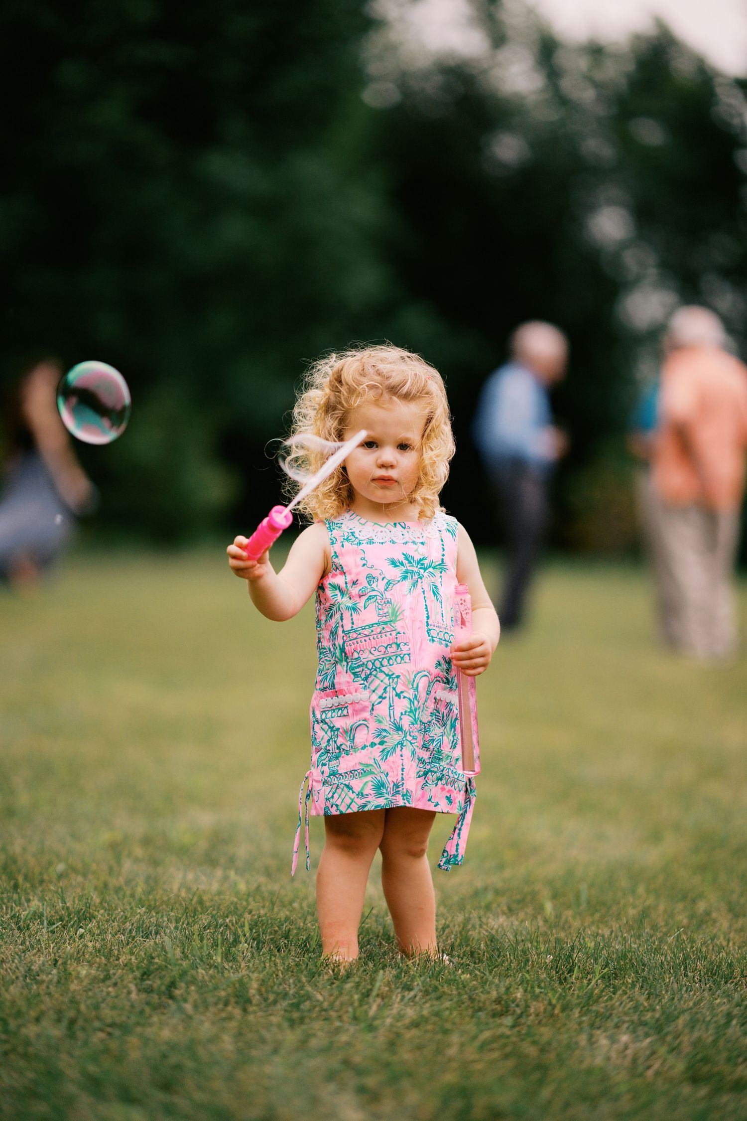 A toddler in a floral dress plays with bubbles on a grassy lawn.