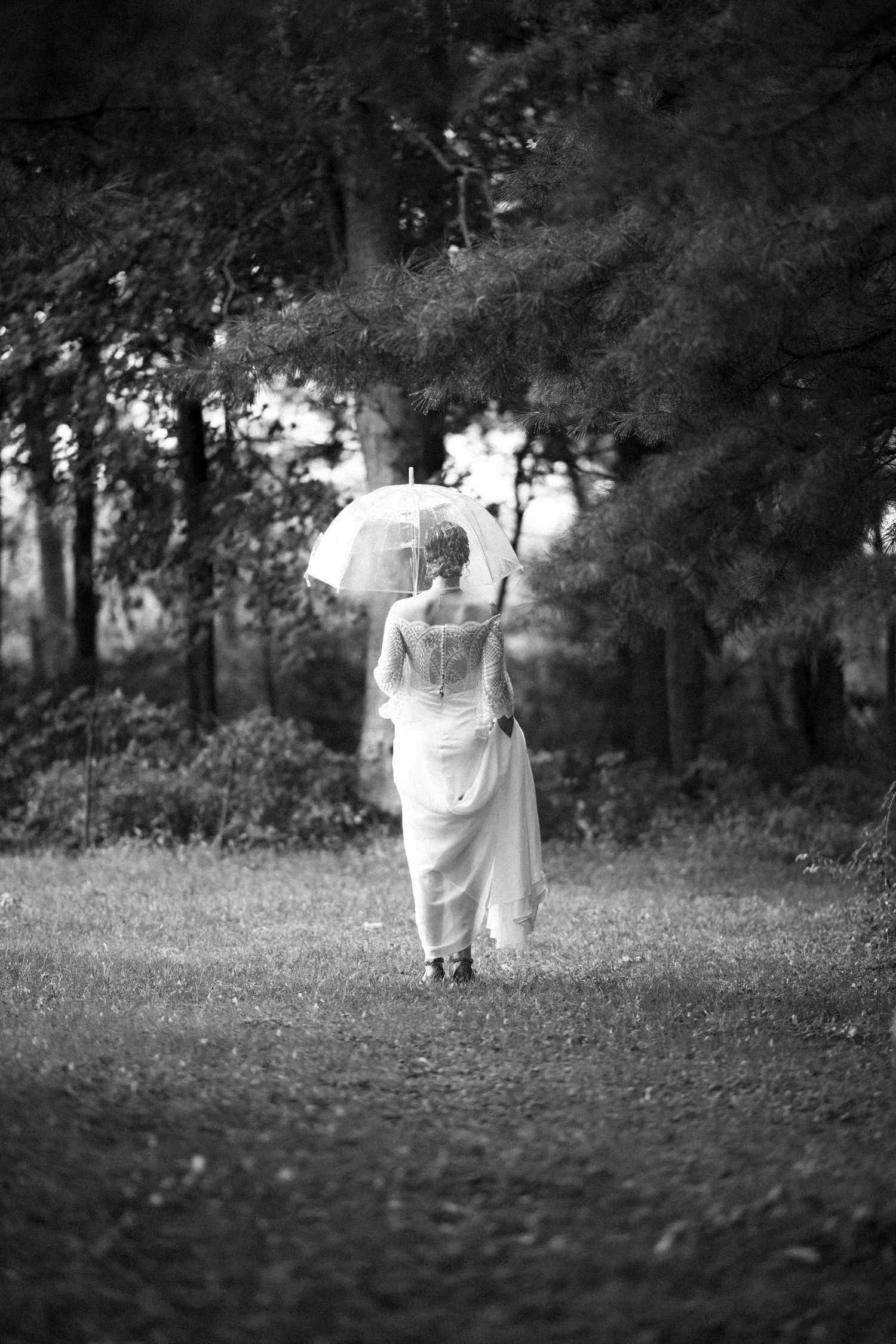 Black and white image of a bride walking through a wooded path holding a parasol.