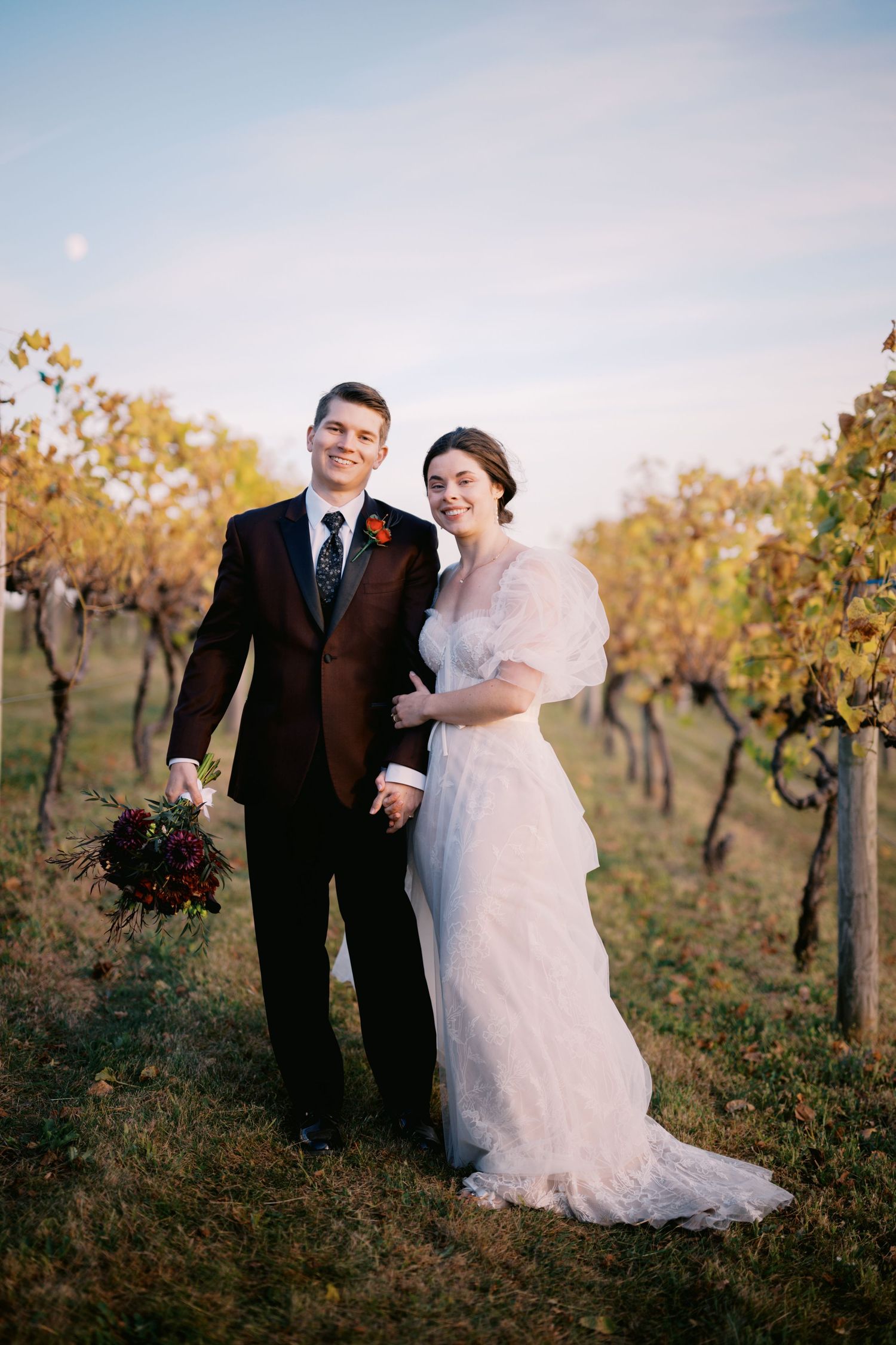 Newlyweds pose together in autumn vineyard with burgundy bouquet at sunset.