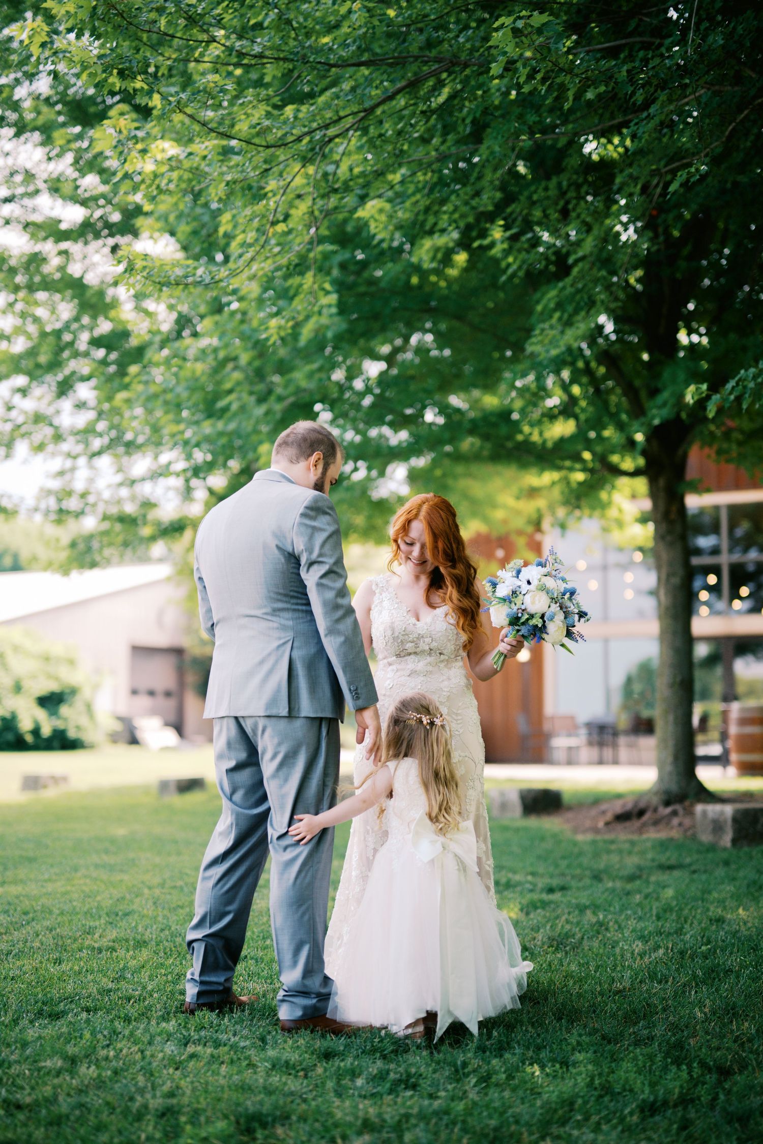 A wedding couple and young child share a tender moment on a lush green lawn surrounded by trees.