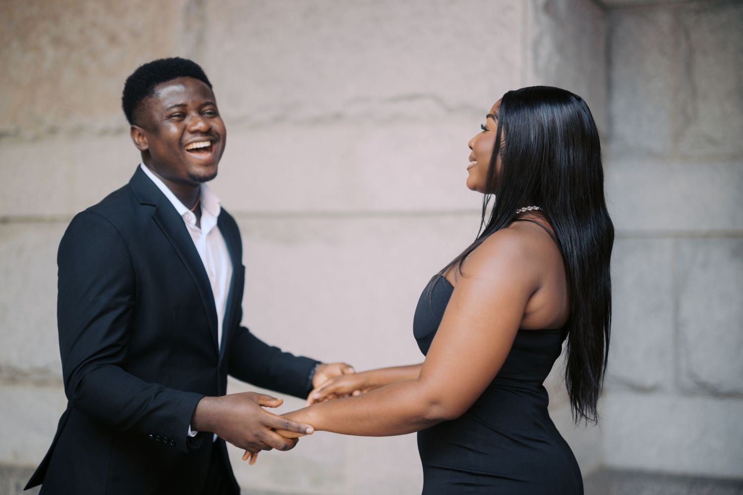A joyful couple holding hands and laughing together against a stone wall backdrop.