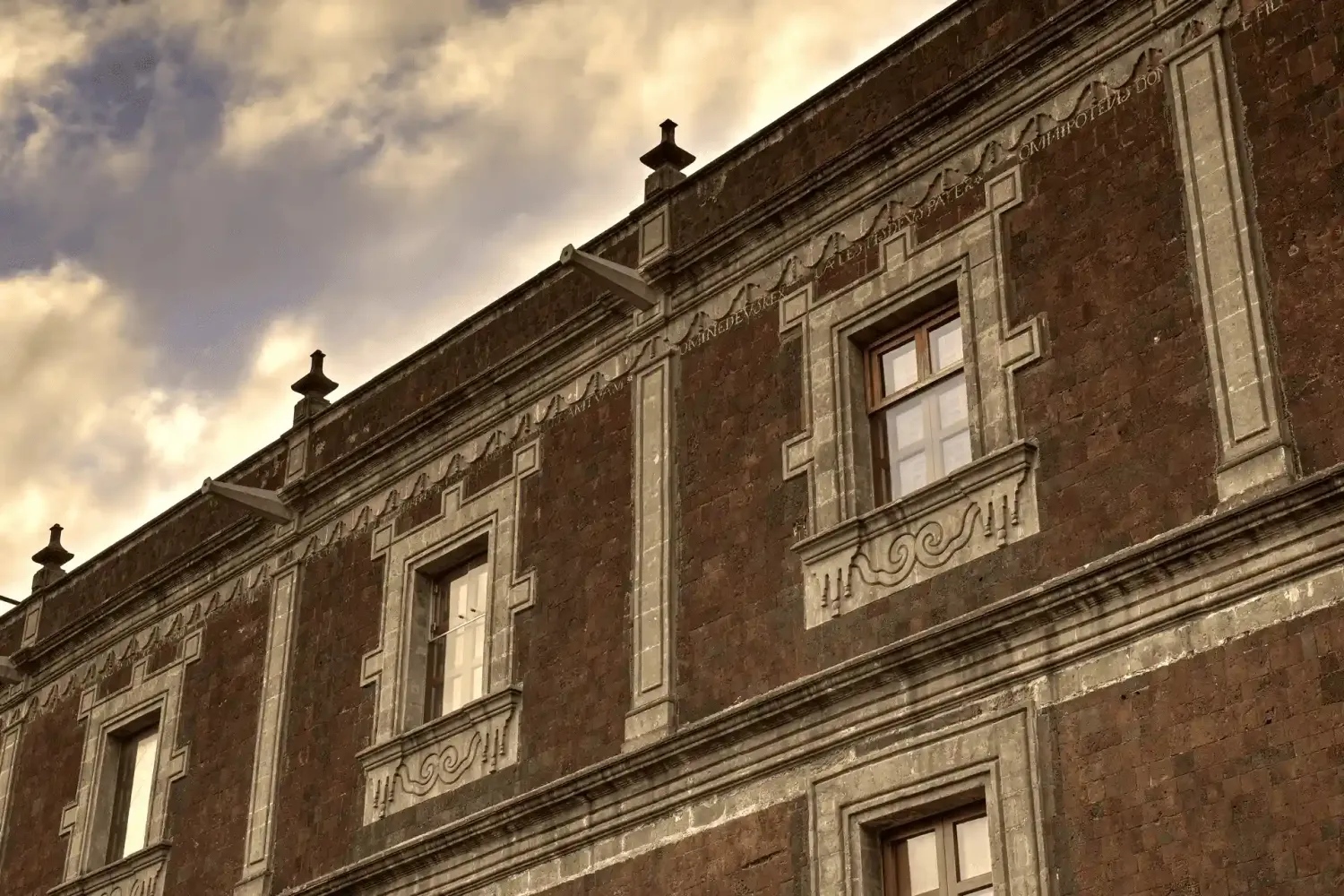 Historic baroque building facade with ornate stone carvings, arched windows, and dramatic cloudy sky in sepia tone.