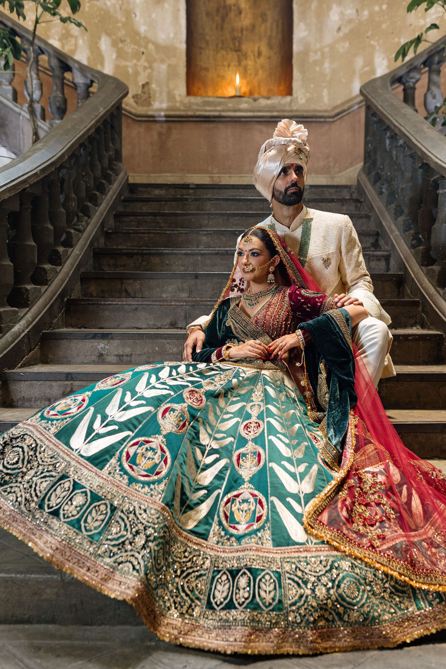 Indian bride in teal lehenga and groom in ivory sherwani pose on ornate staircase in traditional wedding attire.