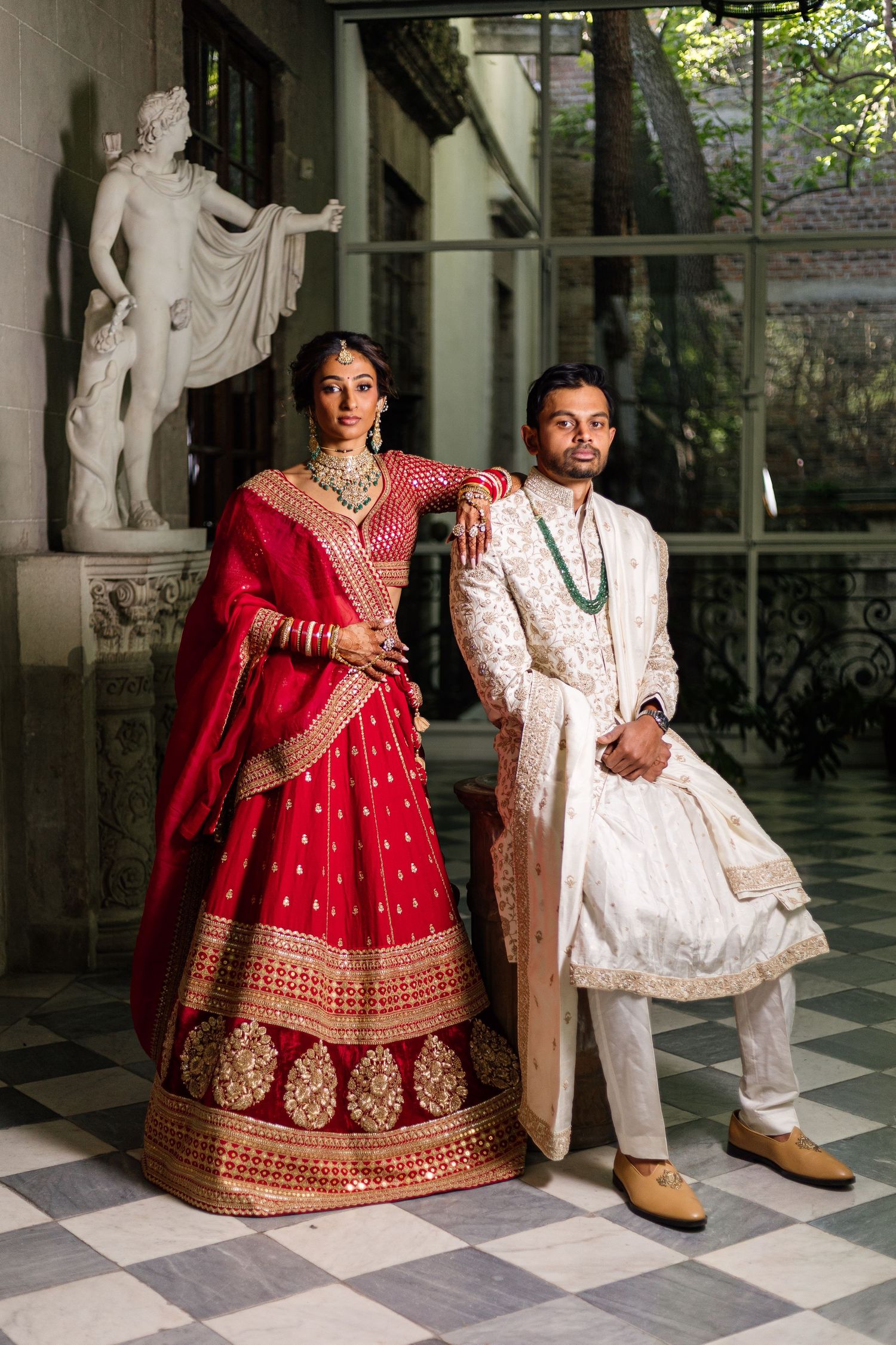 Indian bride in red lehenga and groom in white sherwani pose elegantly before a marble statue.