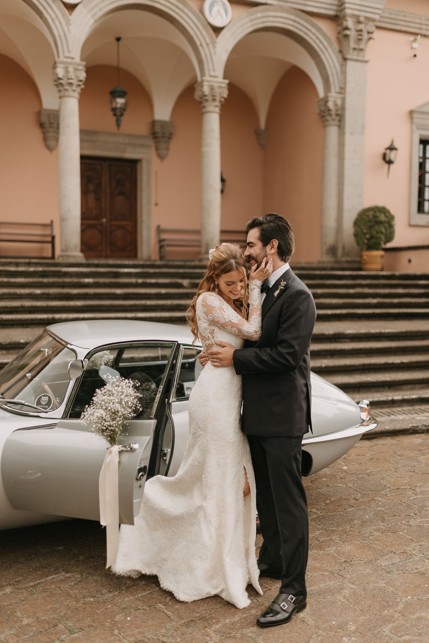 Bride and groom embracing beside a vintage classic car outside an elegant Italian villa on their wedding day.