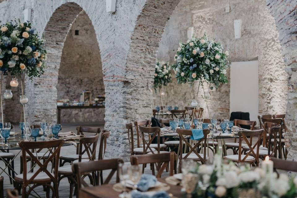 Elegant rustic wedding reception in a stone-arched venue with wooden chairs, blue accents, and tall floral centerpieces.