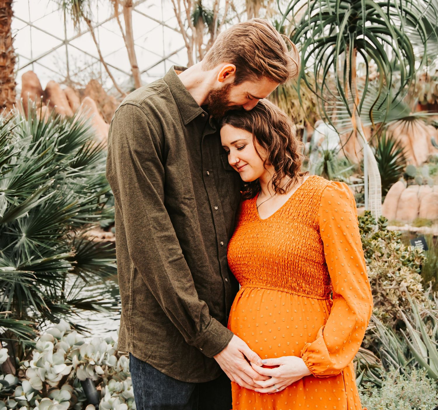 Couple sharing tender moment in lush greenhouse garden setting with orange dress and brown shirt against tropical foliage.