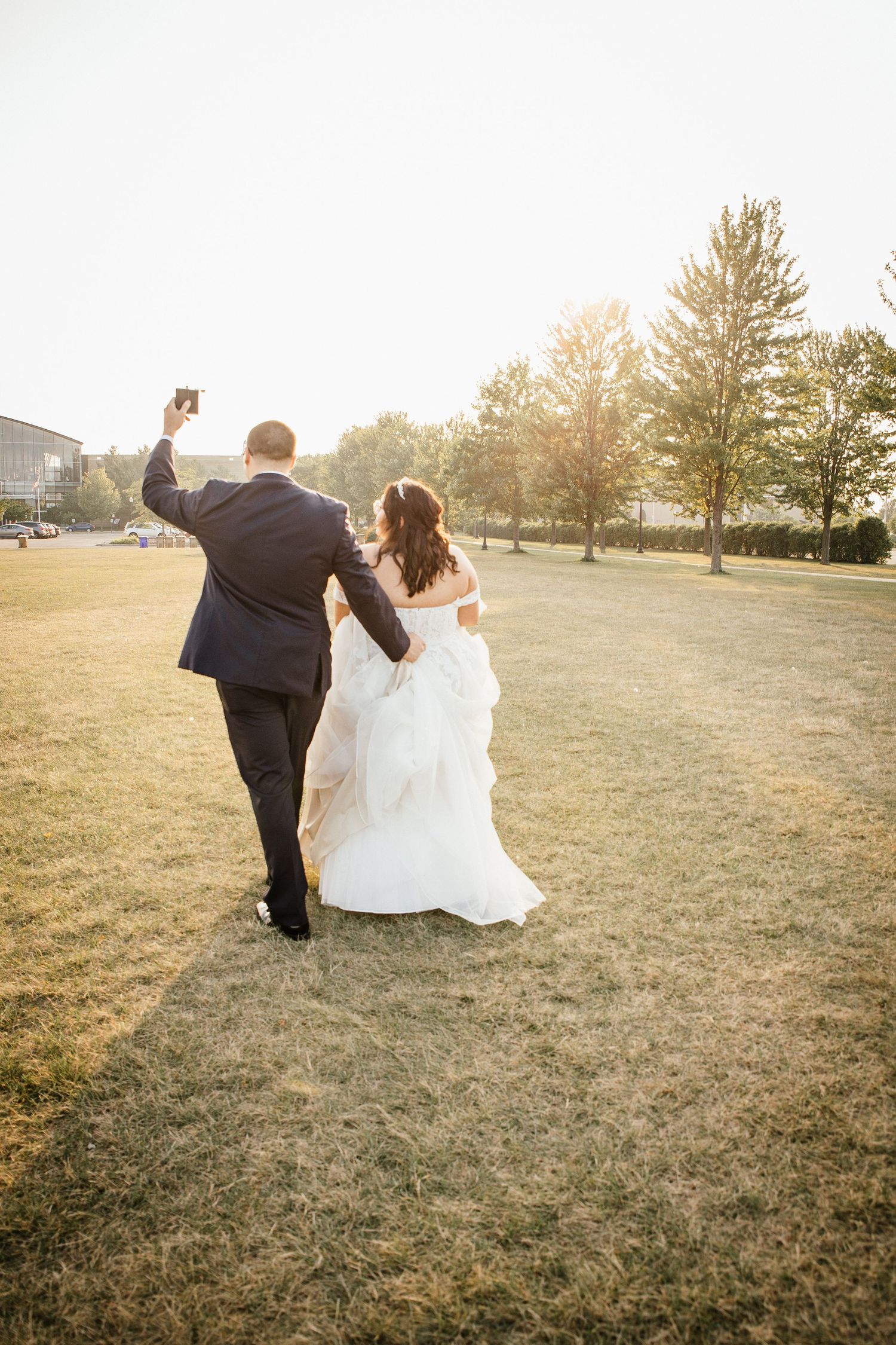A bride and groom walk arm in arm across a sunlit lawn during golden hour at their wedding celebration.
