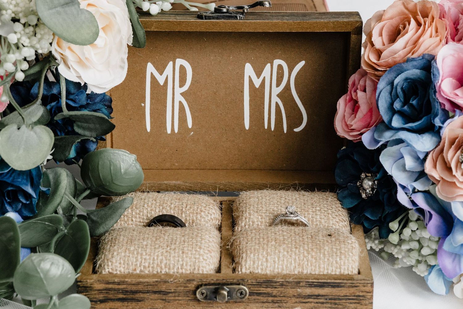 Wedding rings displayed on burlap cushions in rustic wooden box with 'Mr Mrs' text, surrounded by pink and blue flowers.