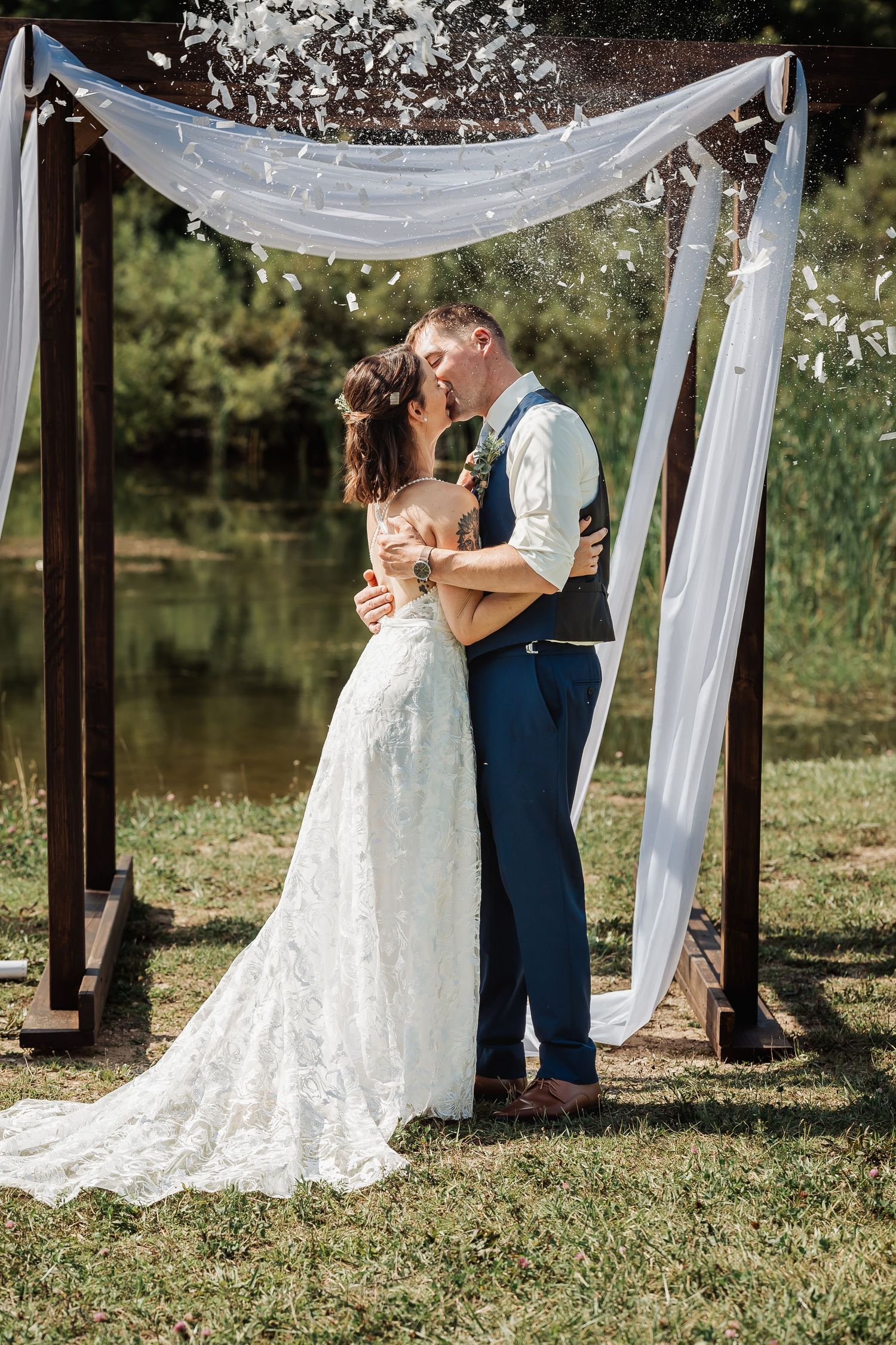 Wedding couple sharing a romantic moment under a white draped arch by a peaceful pond on a sunny day.