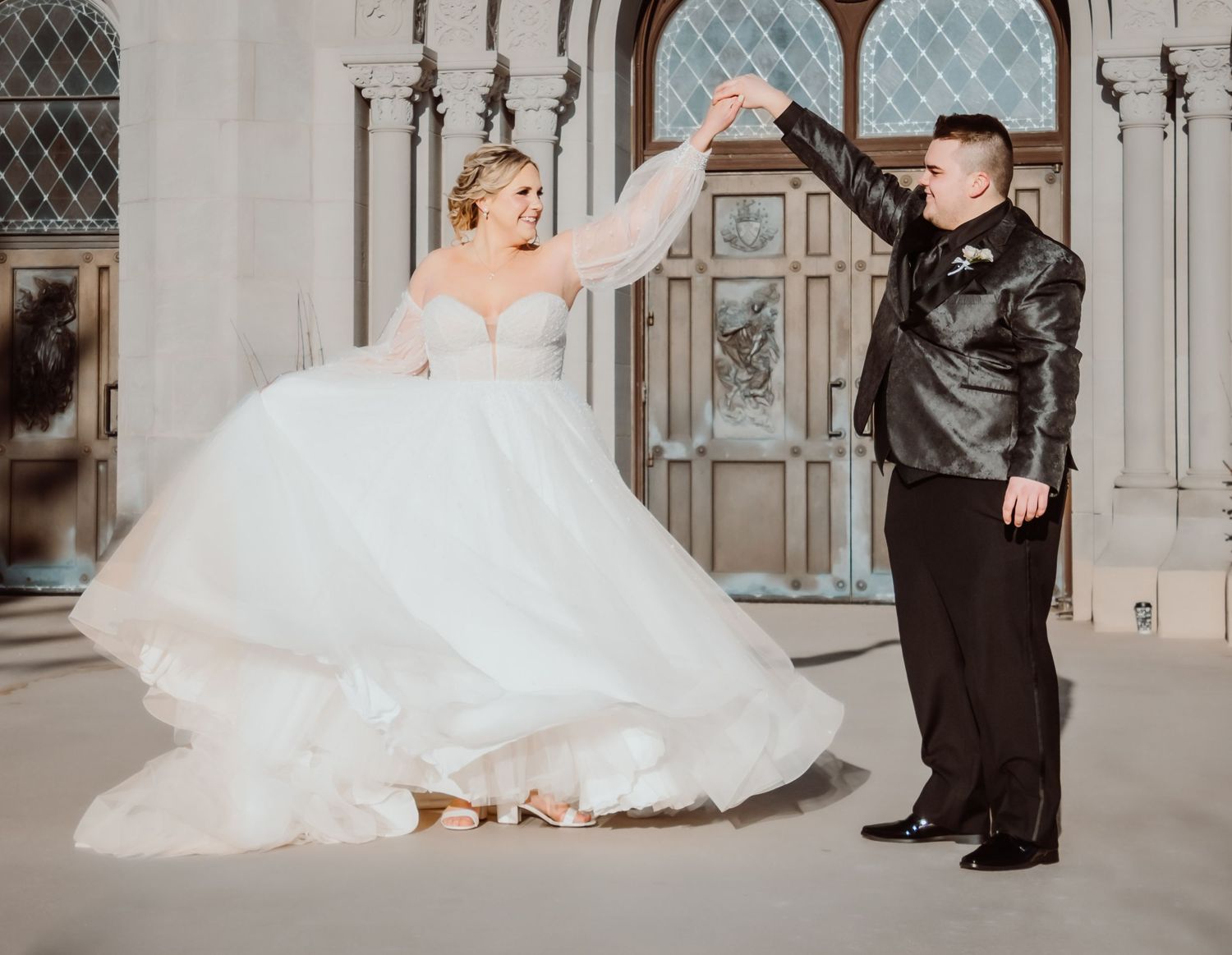 A couple in wedding attire has a playful moment under ornate Gothic architecture with a flowy white ballgown.