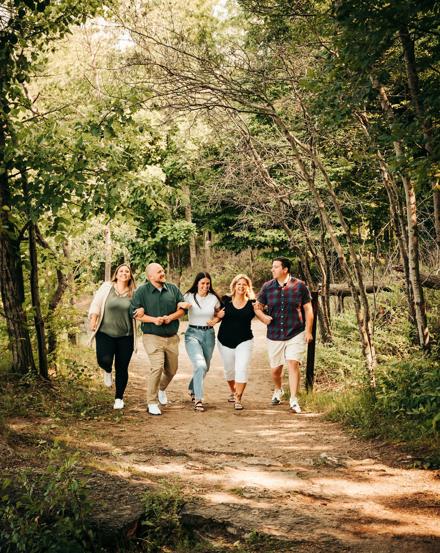 A group walks together along a sunlit wooded trail path during a warm afternoon outdoors.