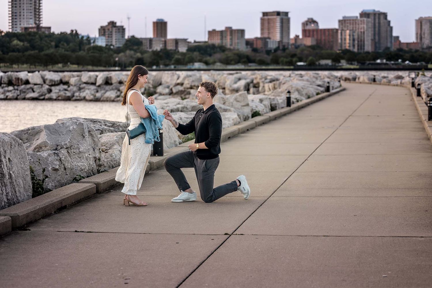 A romantic proposal moment on a waterfront path with city skyline in the background.