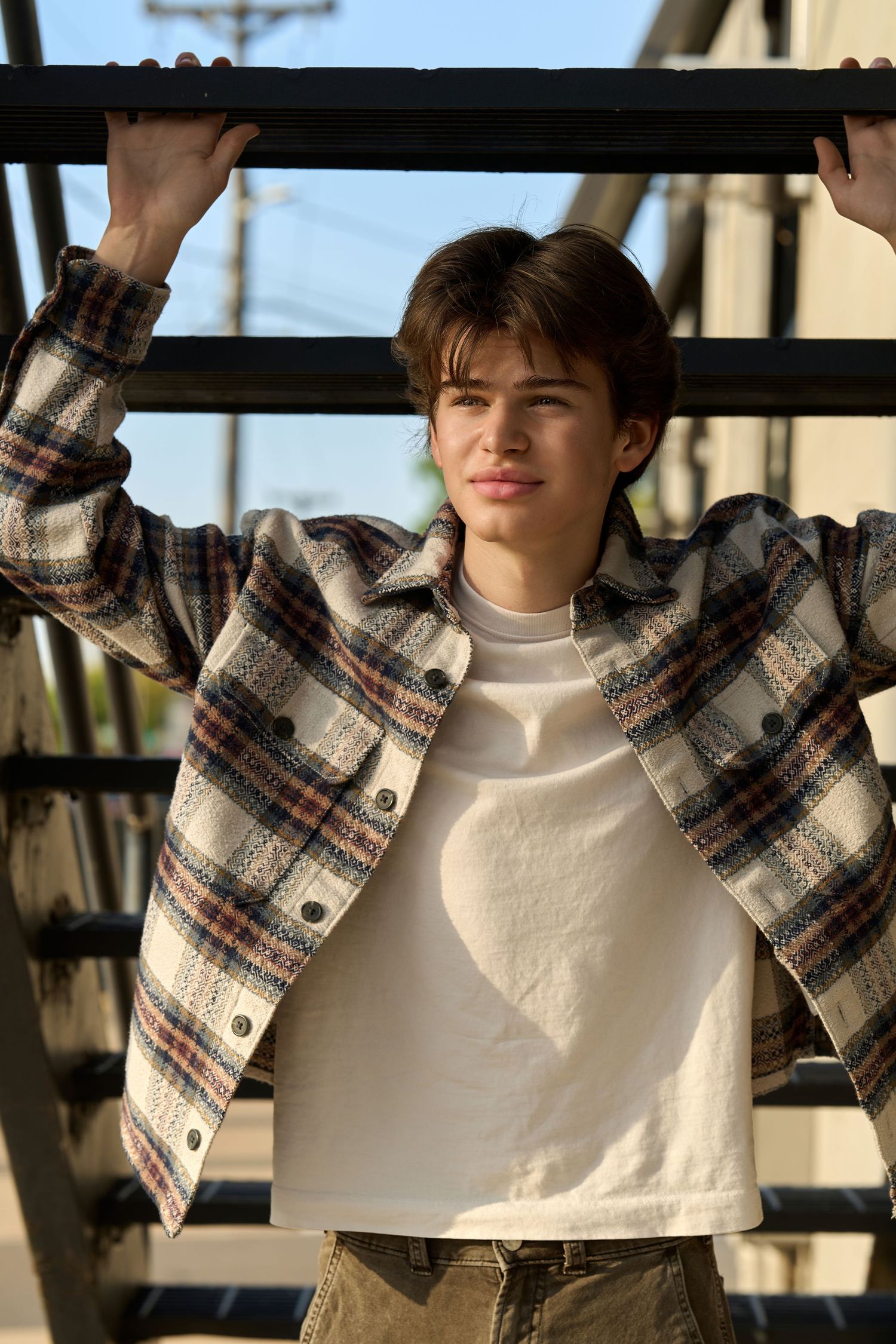 Young man in plaid flannel shirt and white tee holds metal bar outdoors in golden hour sunlight.
