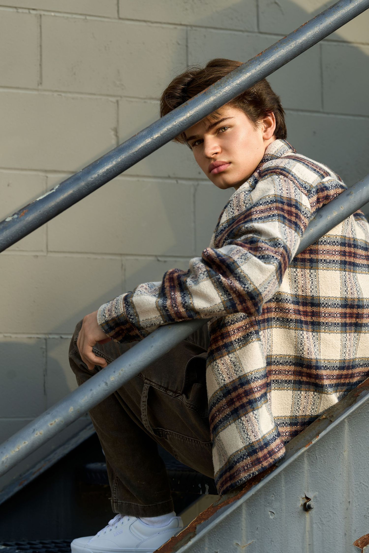 Young man in plaid jacket sitting on metal stairs outdoors, looking at camera with a cool, relaxed expression.
