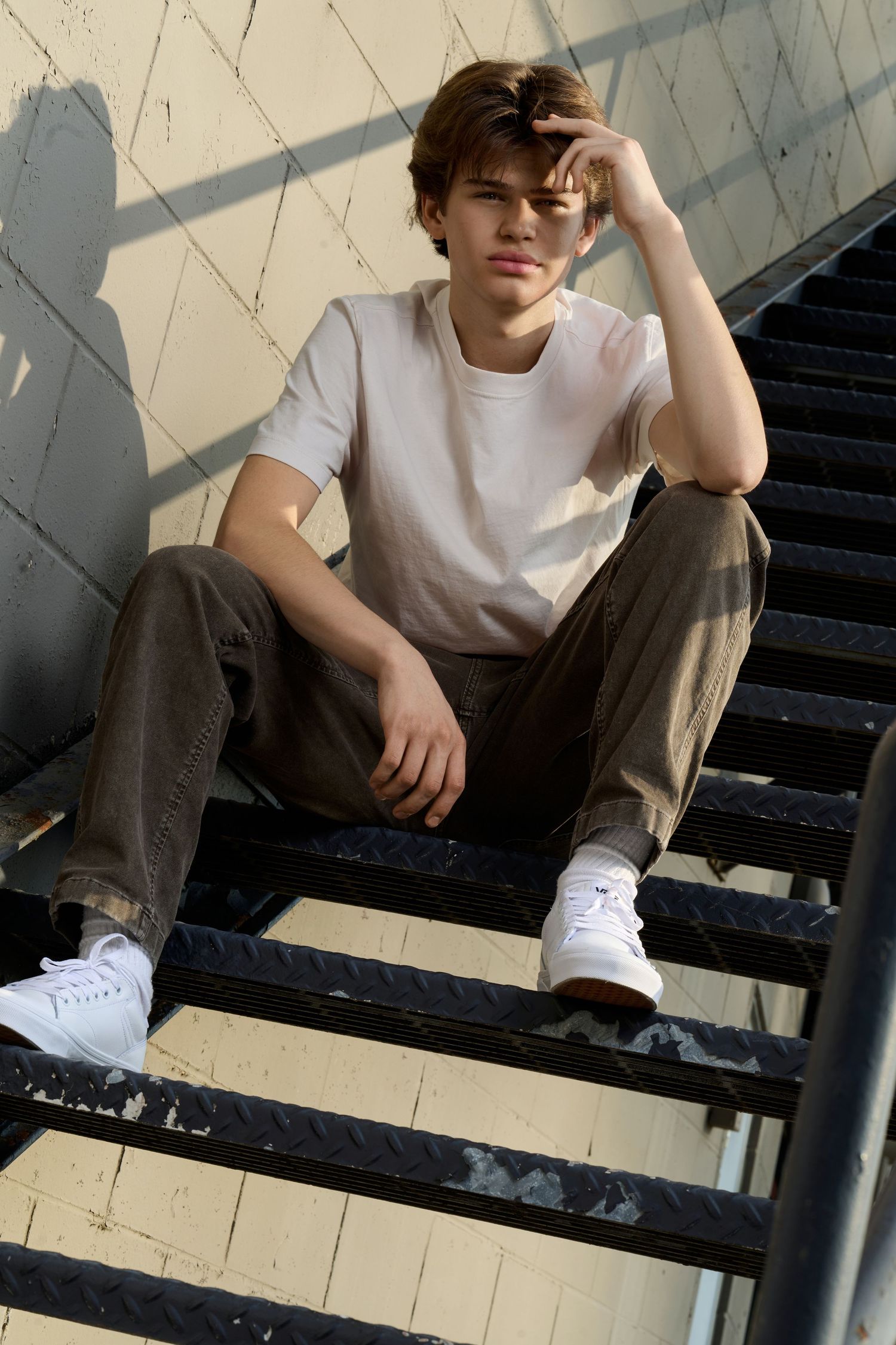 Teen boy in white tee and olive pants sits on metal stairs, resting hand near face, wearing white sneakers.