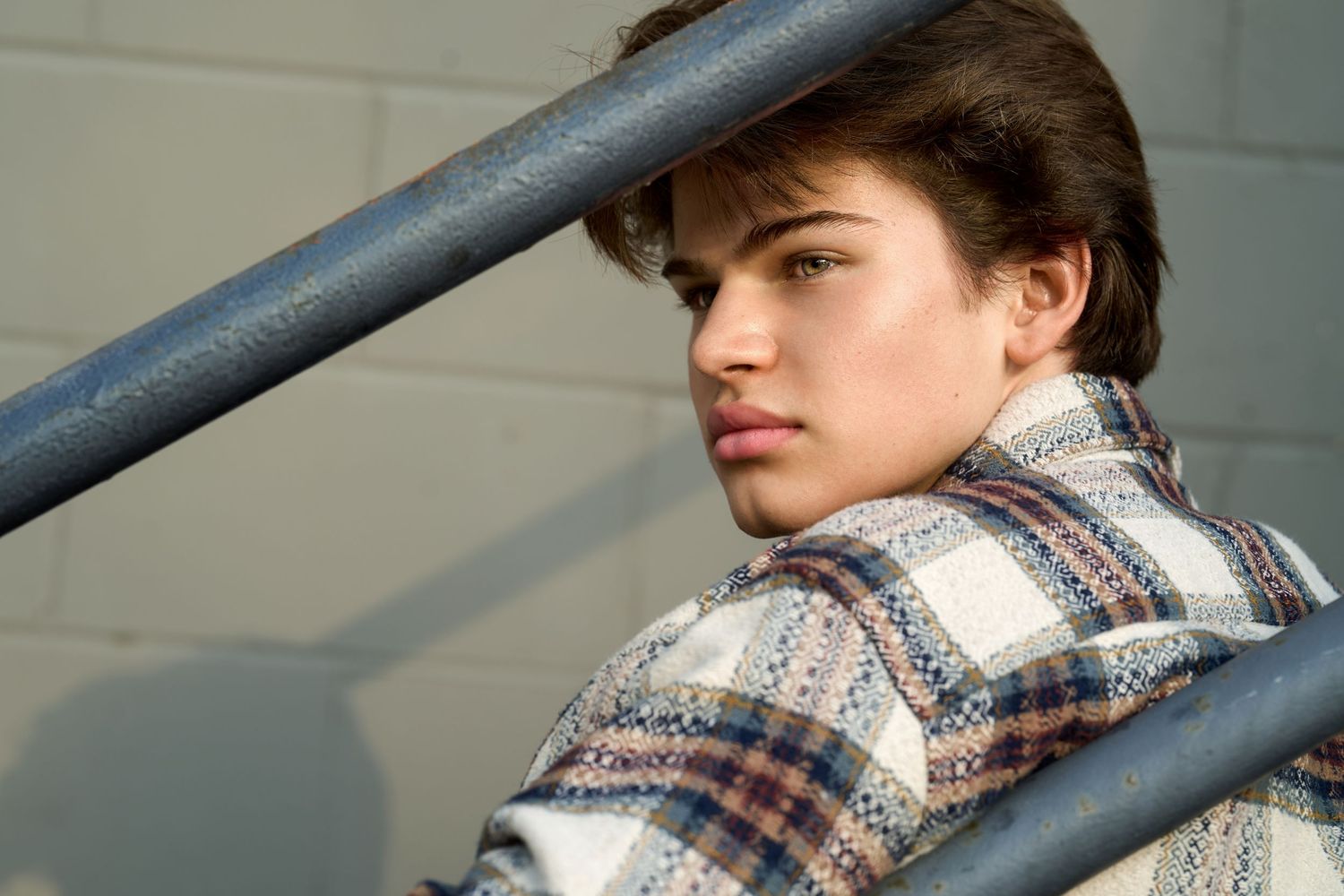 Young man in plaid shirt leaning on metal railing, looking over shoulder with serious expression.