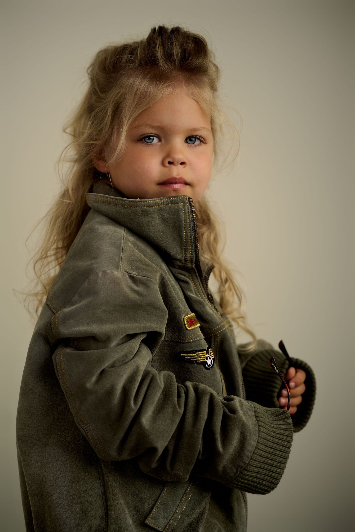 Young girl with long blonde hair wearing an oversized olive green military-style jacket, posing against a light background.