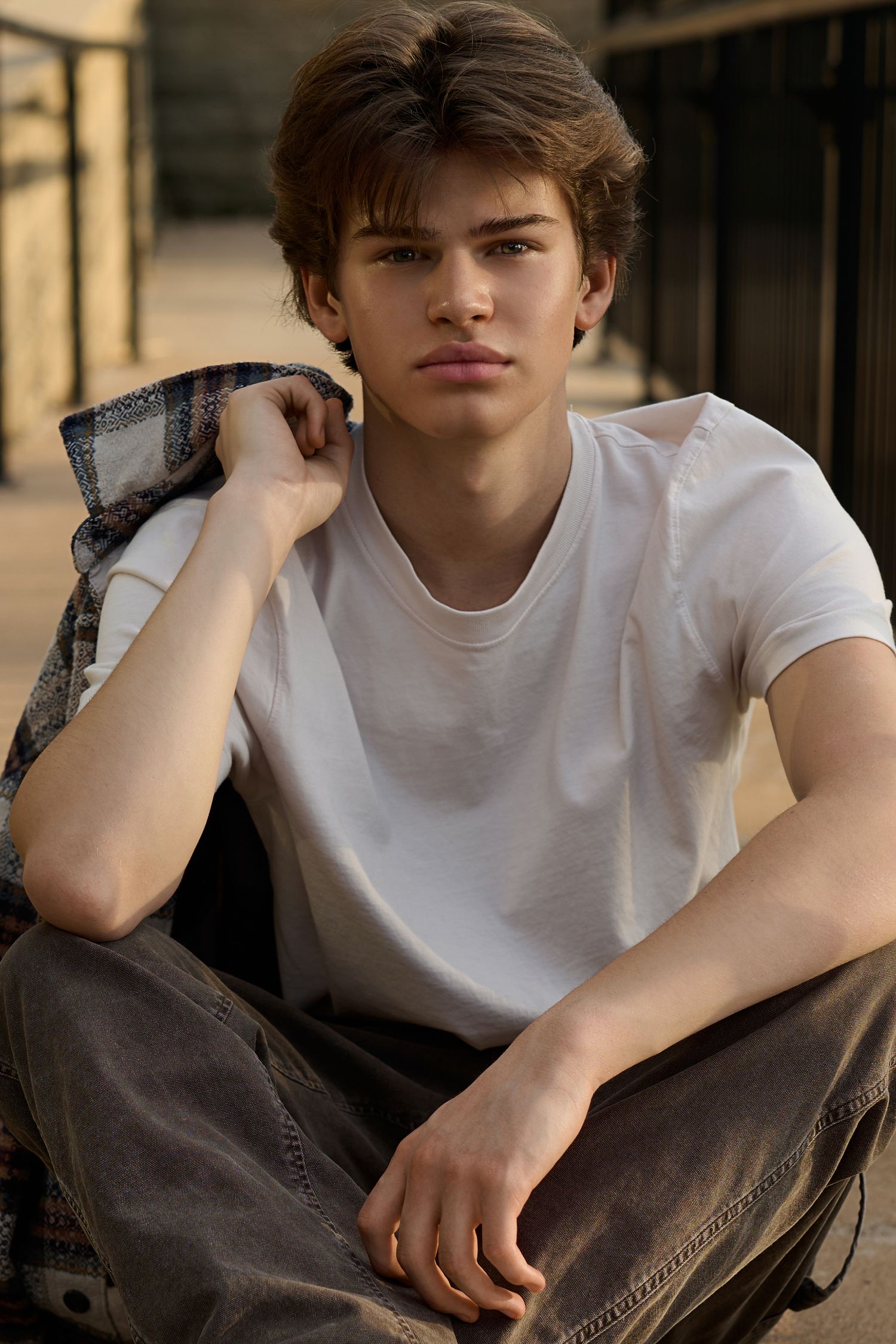 Young man in white t-shirt and flannel jacket sitting outdoors, looking at camera with a calm expression.