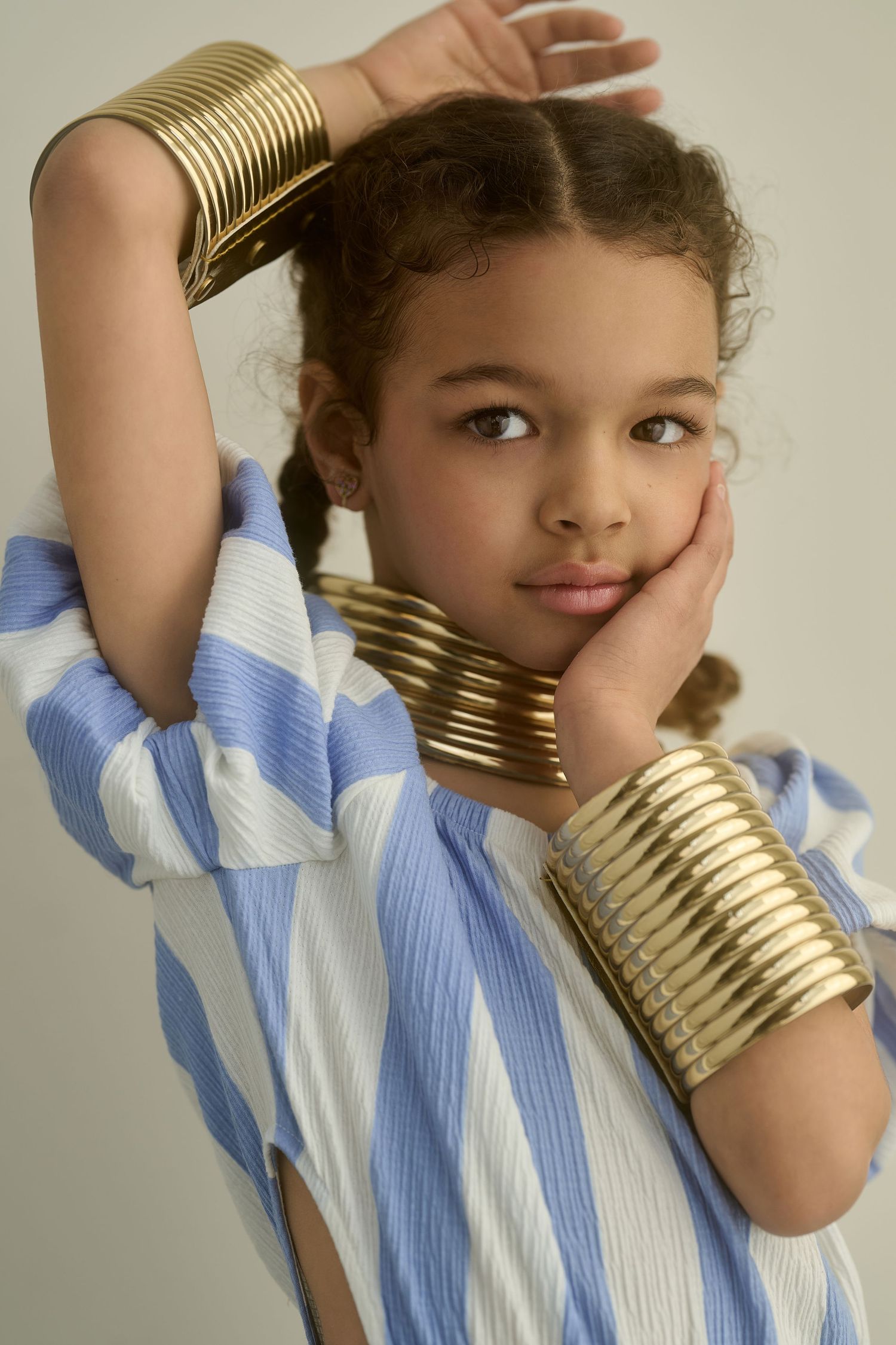 Young girl wearing gold cuff bracelets and neck cuff with blue striped shirt, posing with hand on cheek.