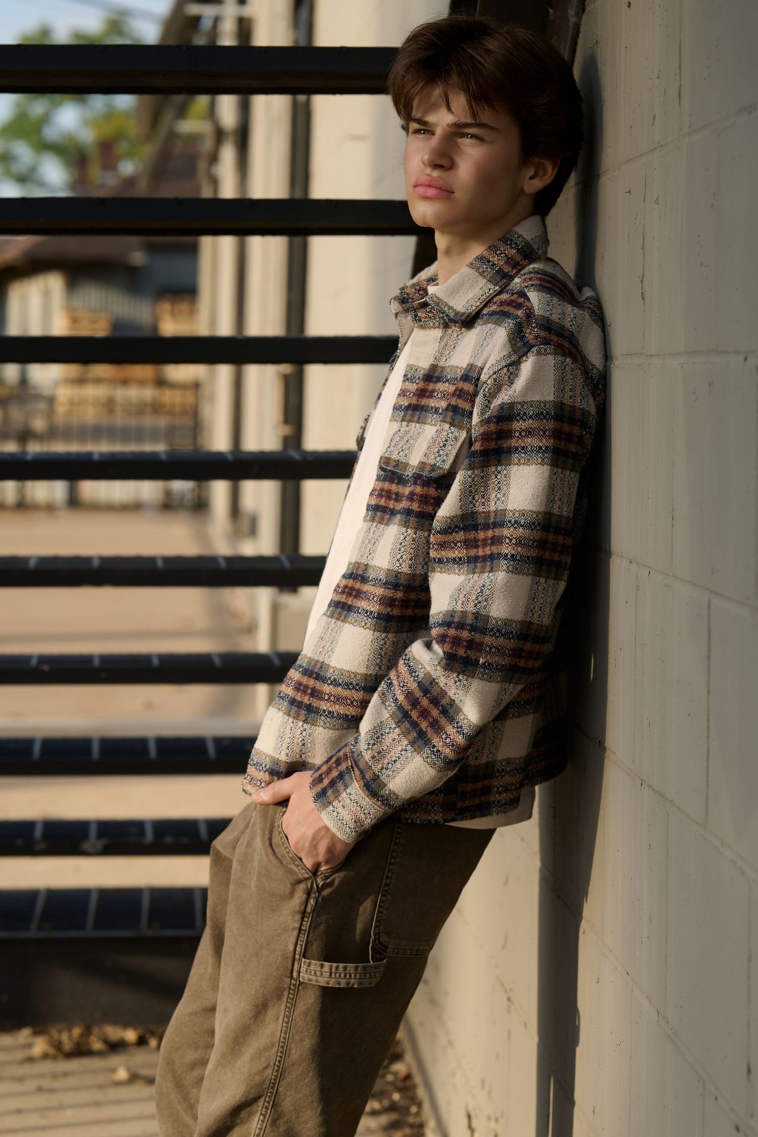 Teen boy in plaid flannel shirt and cargo pants leaning against white brick wall outdoors.