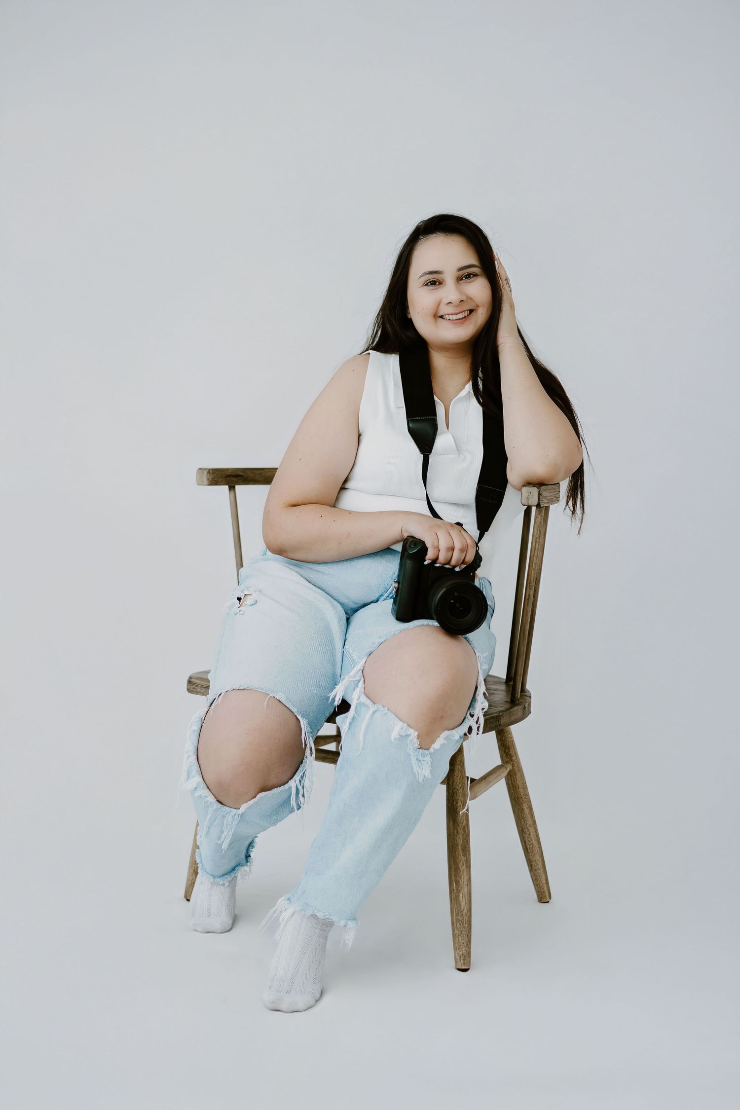Someone wearing a white sleeveless top and ripped light blue jeans sits casually on a wooden chair against a white backdrop.