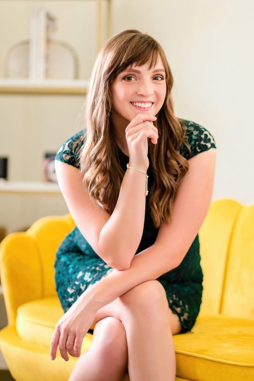 A person in a green lace dress sits on a bright yellow couch with hand resting on chin and smiling warmly.