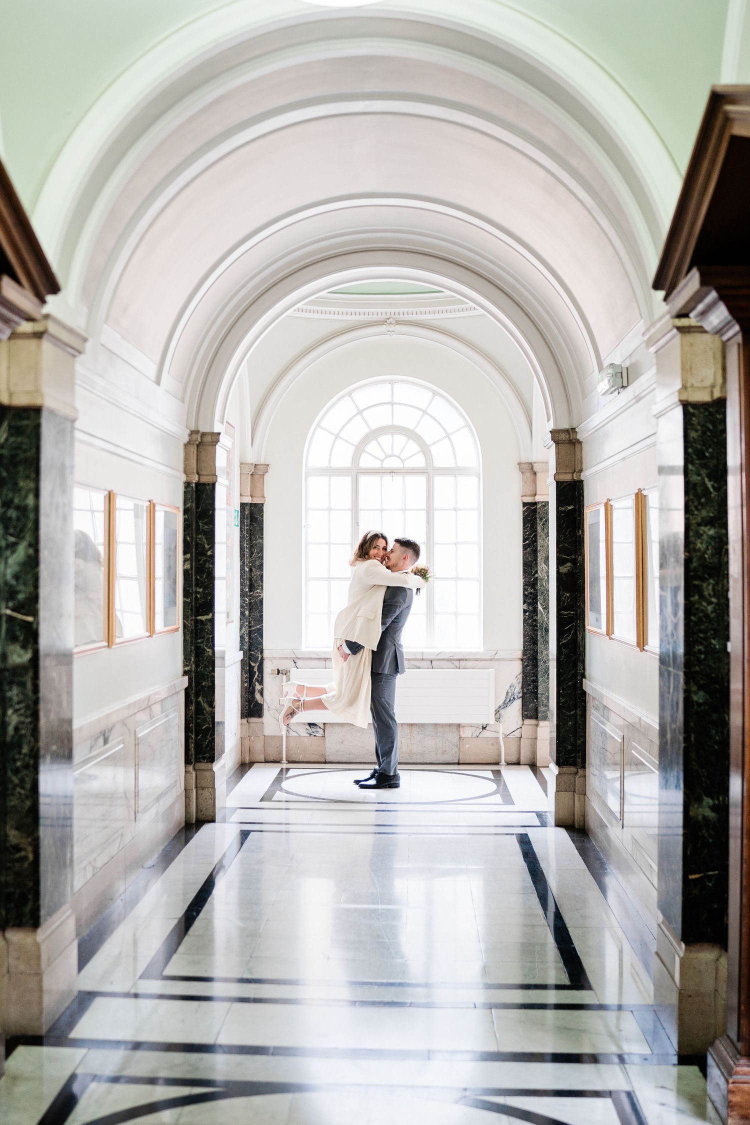Inside Islington Town Hall, the groom lifts the bride in a joyful embrace just after their wedding ceremony, surrounded by elegant interiors