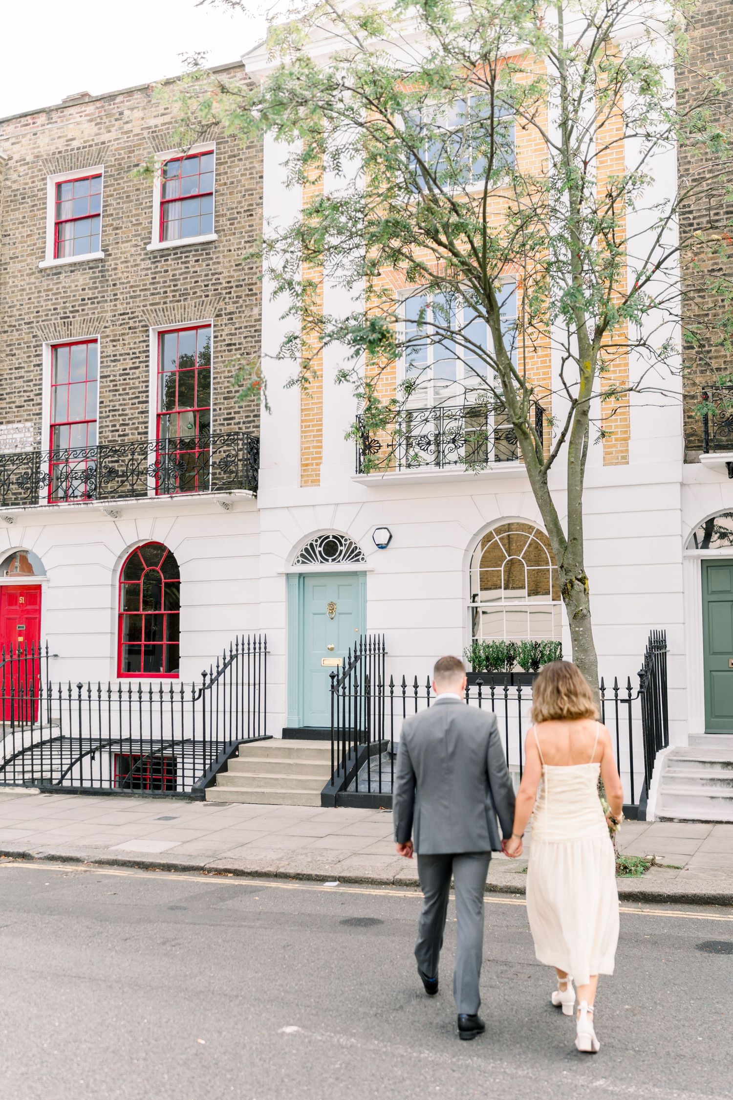 Bride & groom walking hand in hand in front of colourful urban houses near Islington Town Hall in Central London, just after saying 'I do'.
