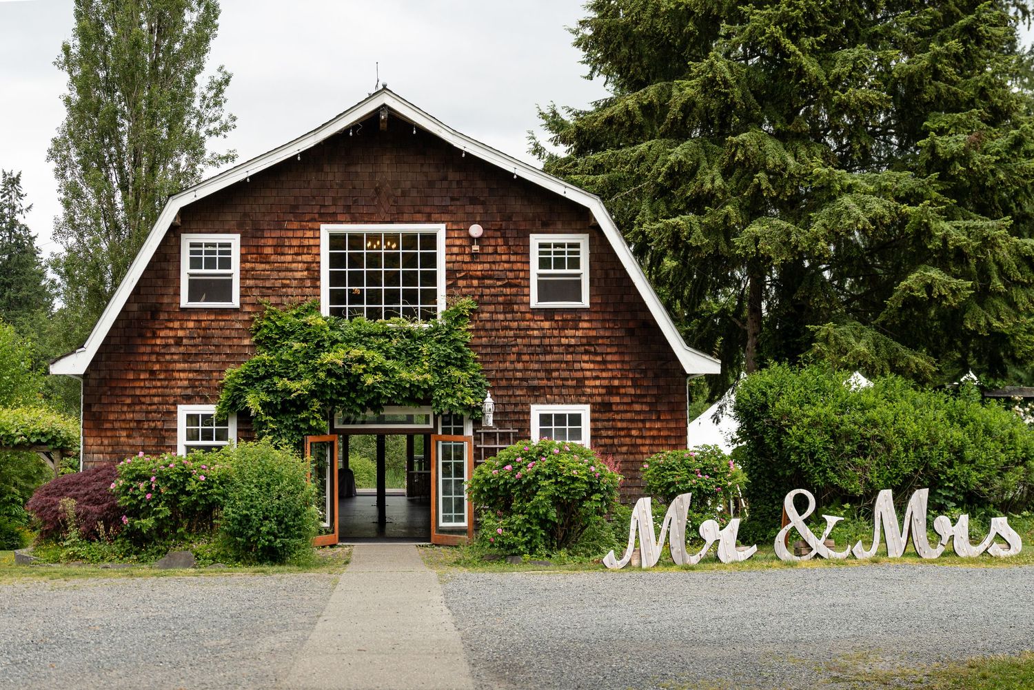 A rustic brown barn venue with large windows and gravel driveway surrounded by green trees and landscaping. Captured by Savvy Photography