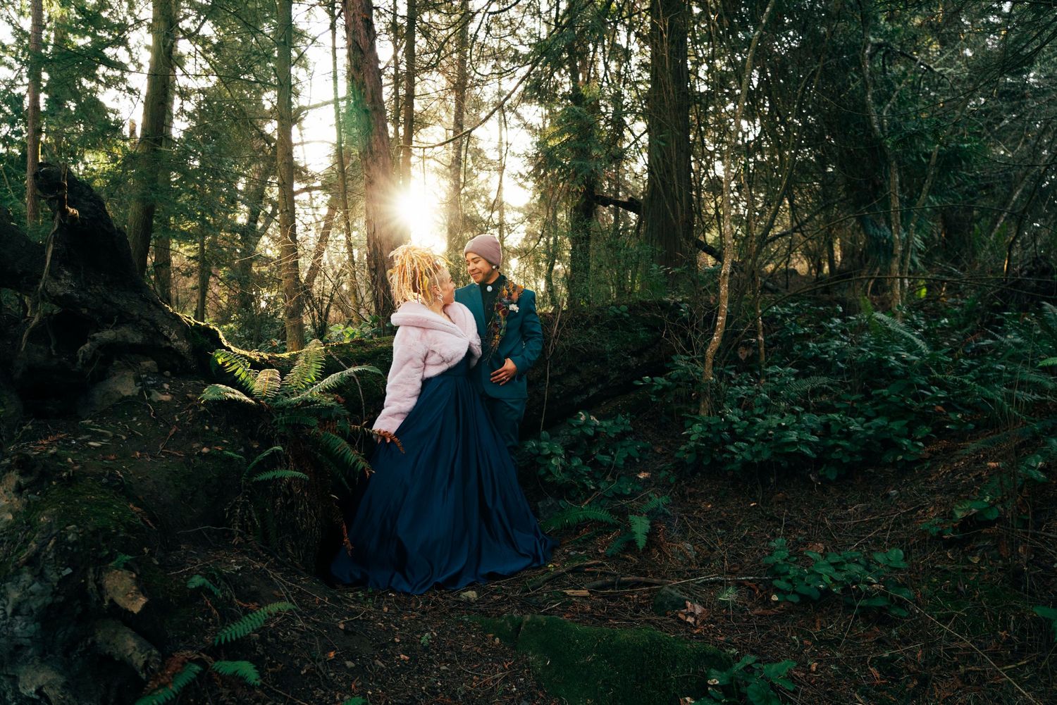 A couple walks through a sunlit forest path with tall trees casting long shadows during golden hour. Captured by Savvy Photography