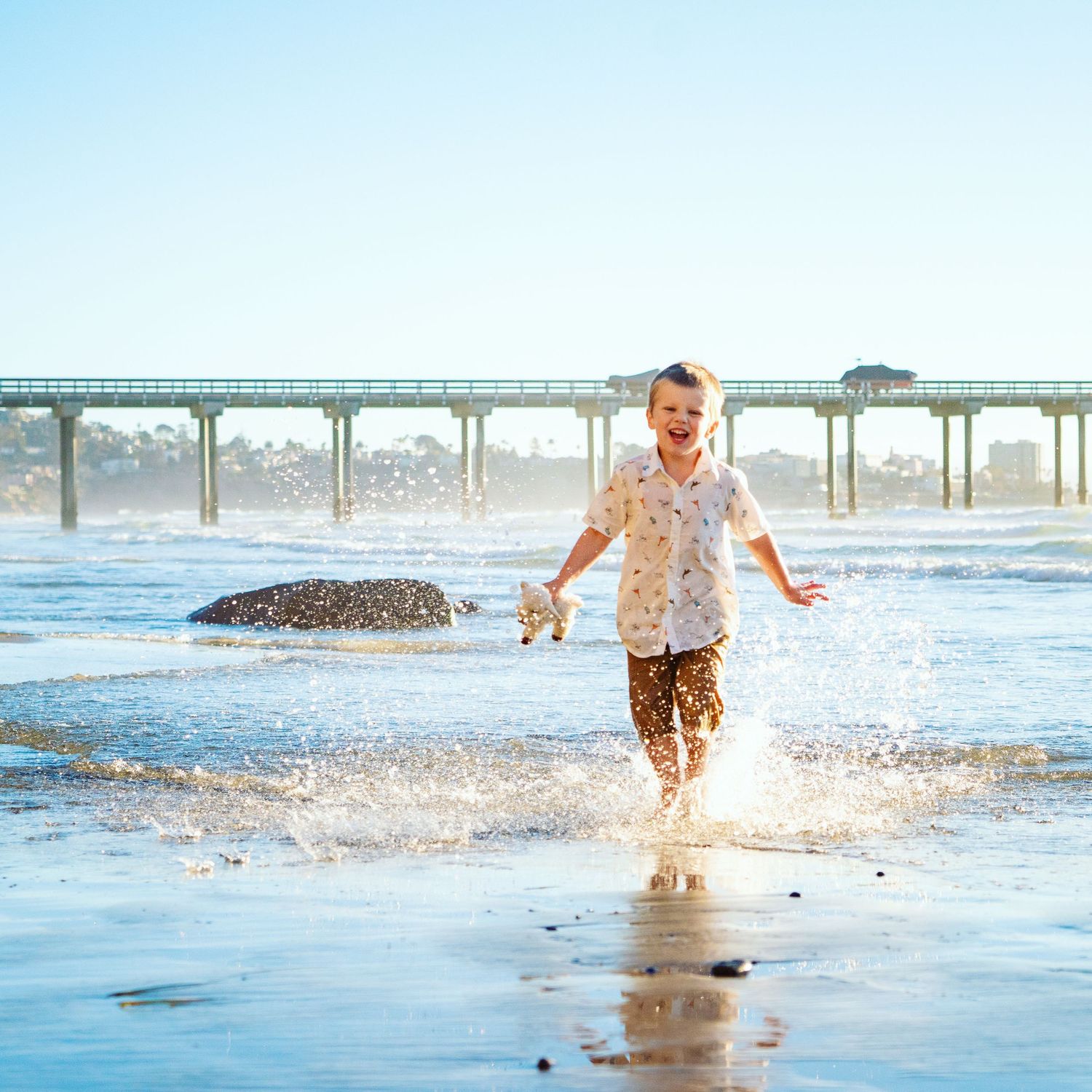 Joyful young boy splashing through shallow ocean water at sunset near a long pier with waves and misty atmosphere, captured by Savvy