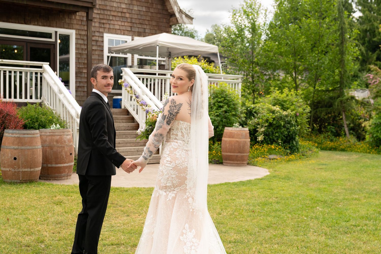 A couple in wedding attire share a romantic moment in front of a brick house with a green lawn. Captured by Savvy Photography.