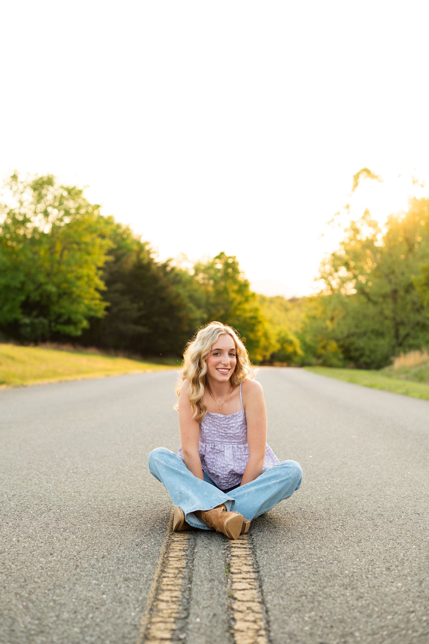 Young blonde woman sitting cross-legged on a road at golden hour, smiling, wearing jeans and a lavender top.