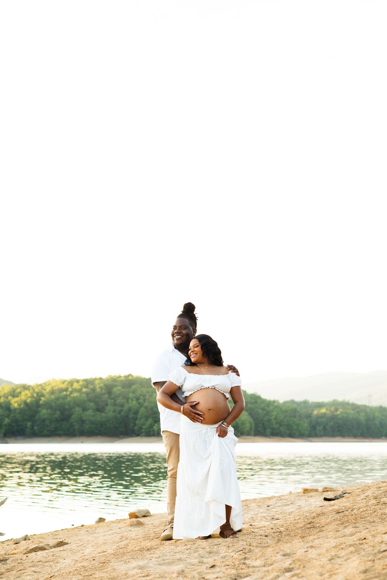 A couple poses by a lakeside, the man embracing a pregnant woman in a white dress during a maternity photoshoot.