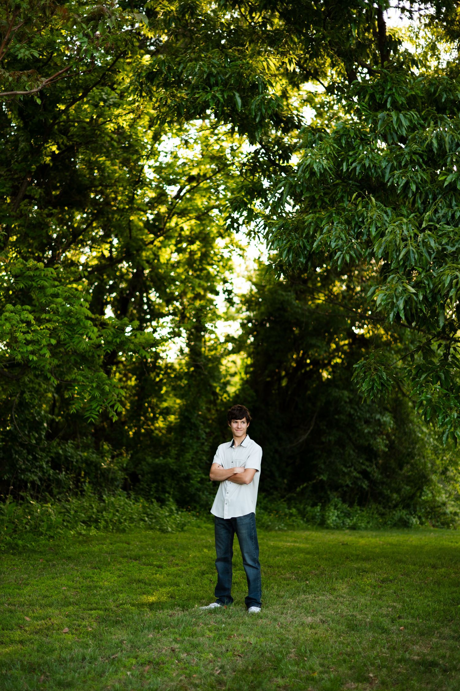 Young man standing in a lush green park with arms crossed, surrounded by tall trees on a sunny day.